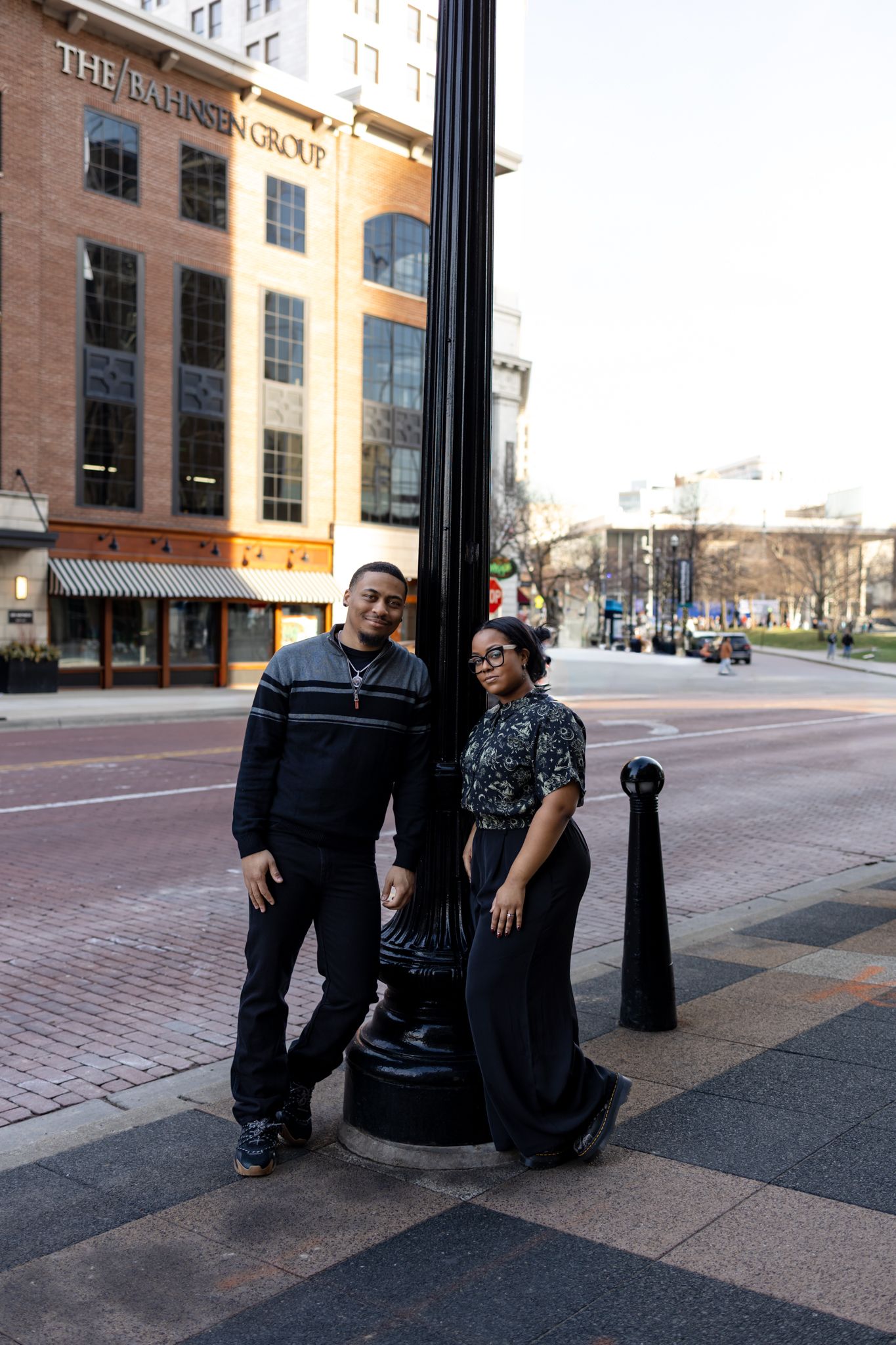 Two people stand together on a brick sidewalk in front of The Boston News Cafe building on a city street.