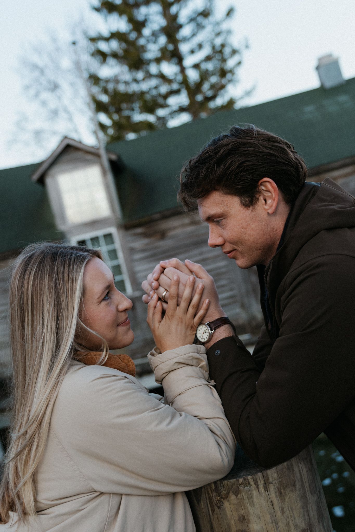 Romantic couple holding hands outdoors near a rustic building, woman wearing engagement ring, man with watch.