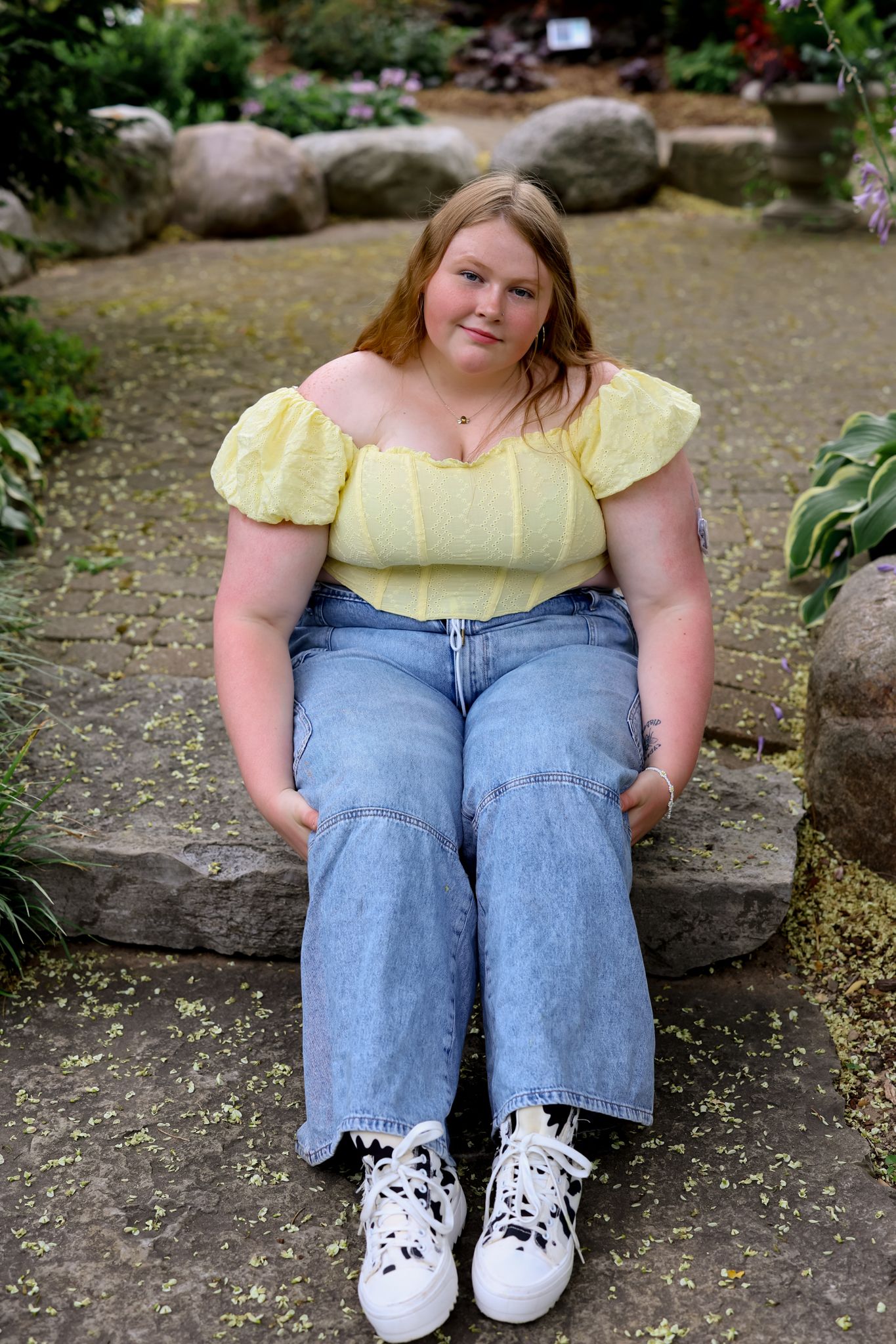 Person in yellow top and jeans sitting on stone ledge outdoors.