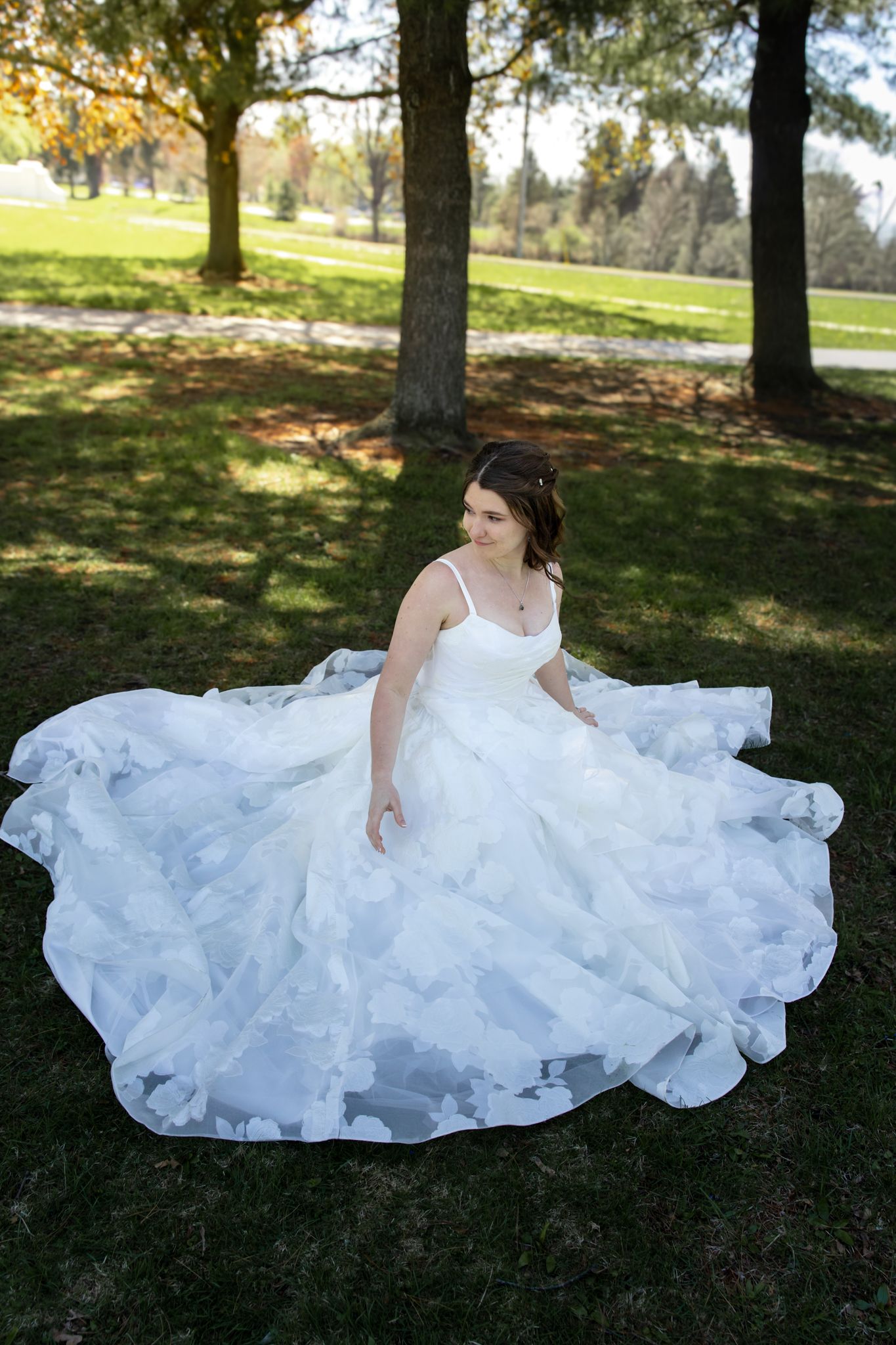 Bride twirls in stunning layered white ball gown with floral details in sunlit park setting.