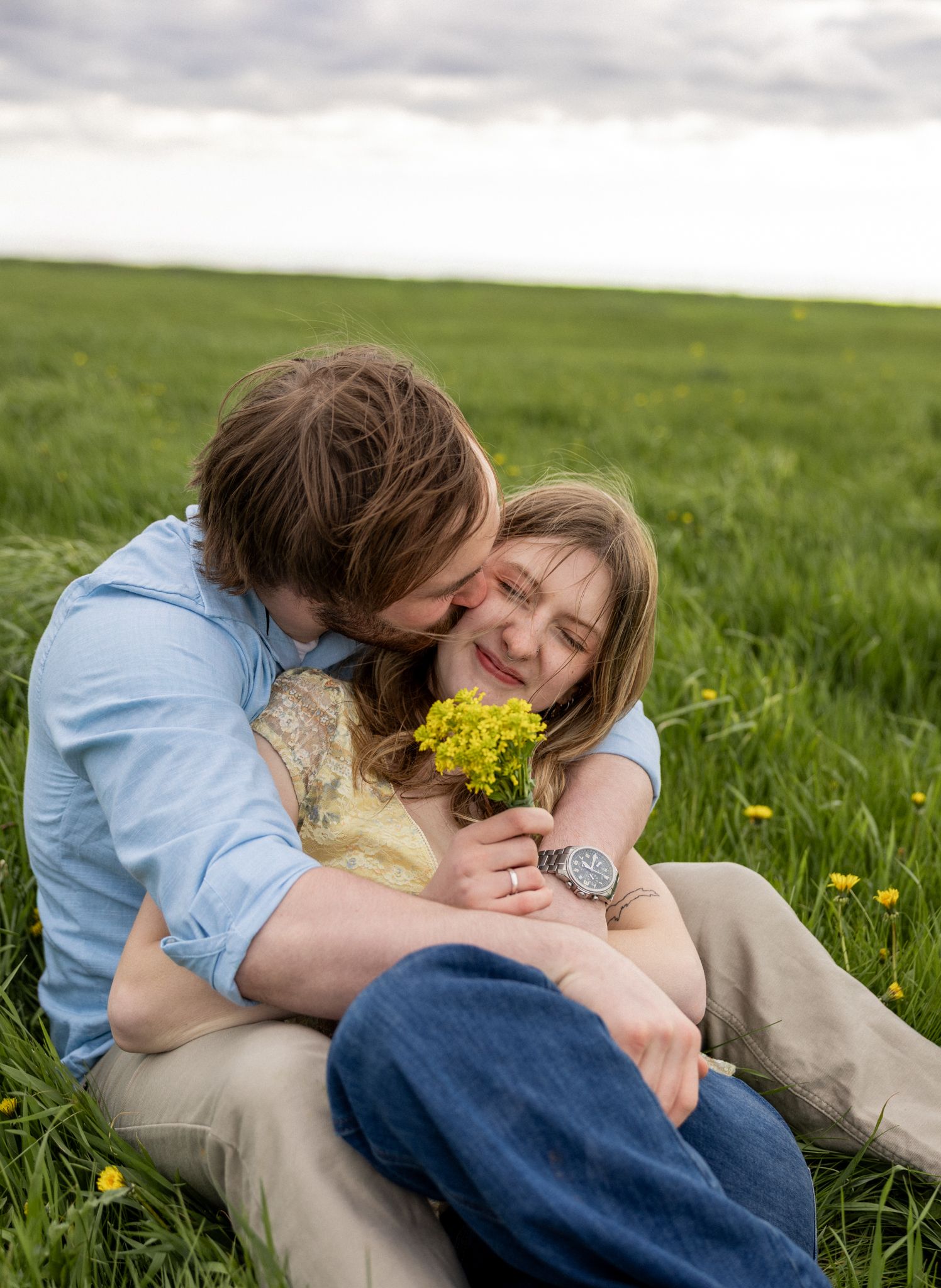 Couple sitting in green field, man kissing woman's cheek as she smiles holding yellow wildflowers.