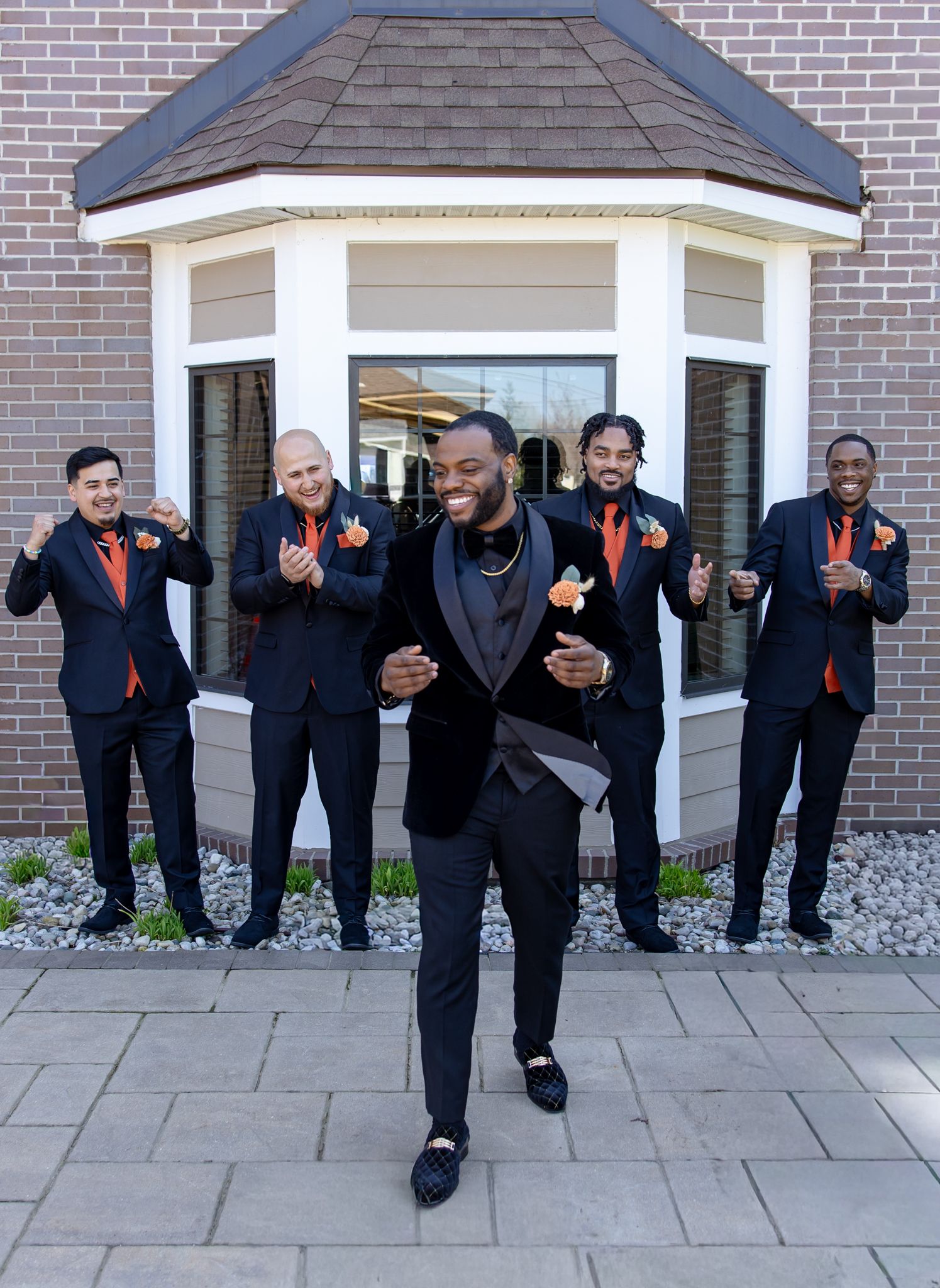 Five groomsmen in black tuxedos with orange boutonnieres pose outside a brick building, with the groom leading confidently.