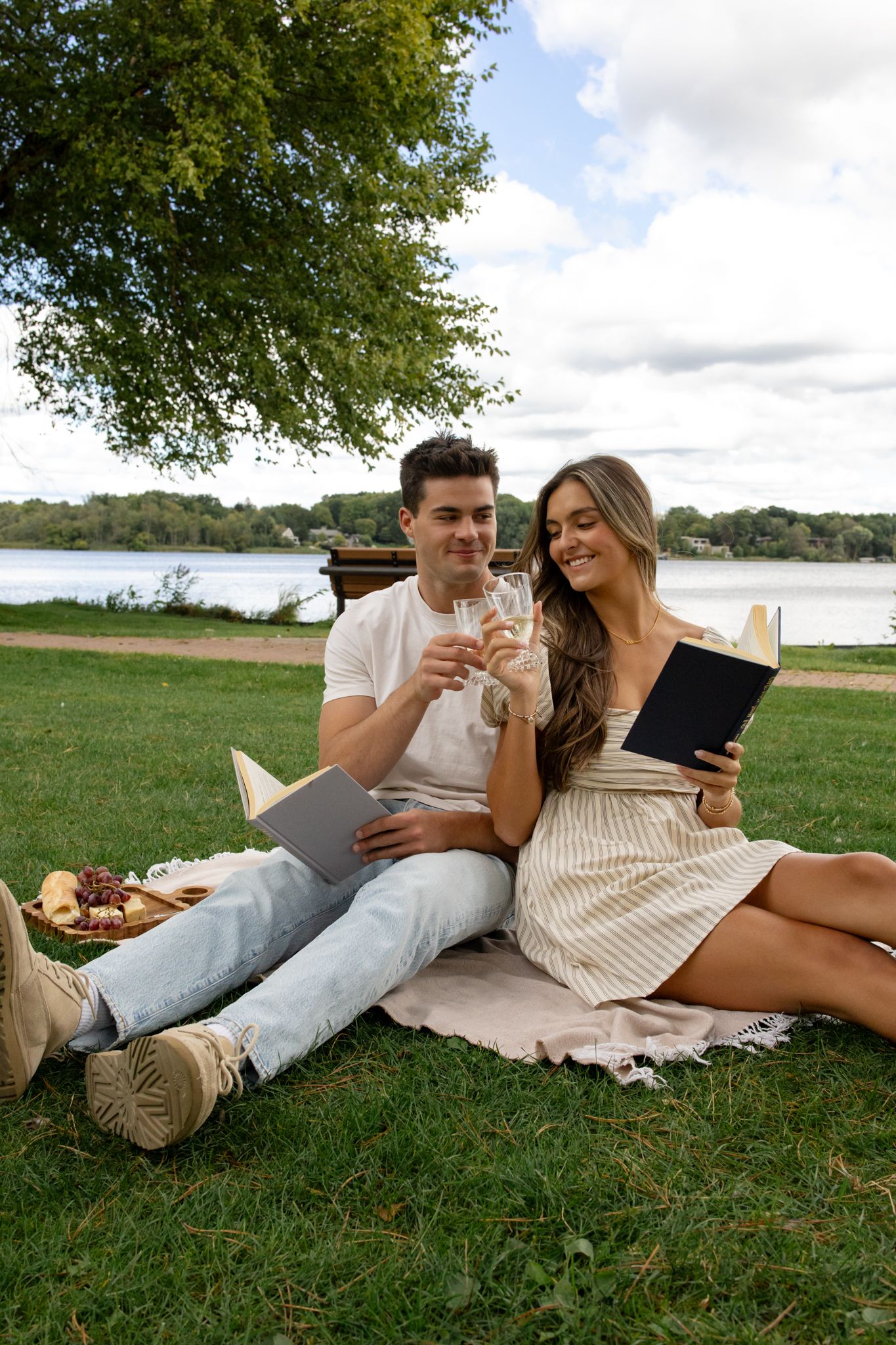 A couple relaxes on a blanket sharing drinks and looking at a tablet by the lake.