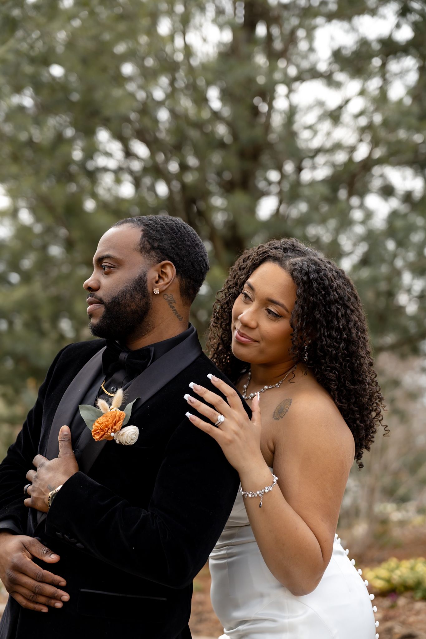 A bride in white leans on her groom in a black velvet tuxedo with an orange boutonniere outdoors.