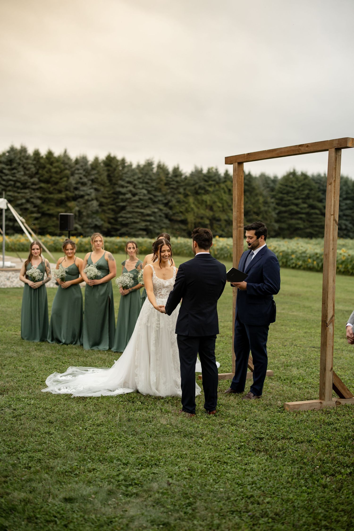 Wedding party gathered under a wooden arch frame in an outdoor ceremony setting with pine trees.