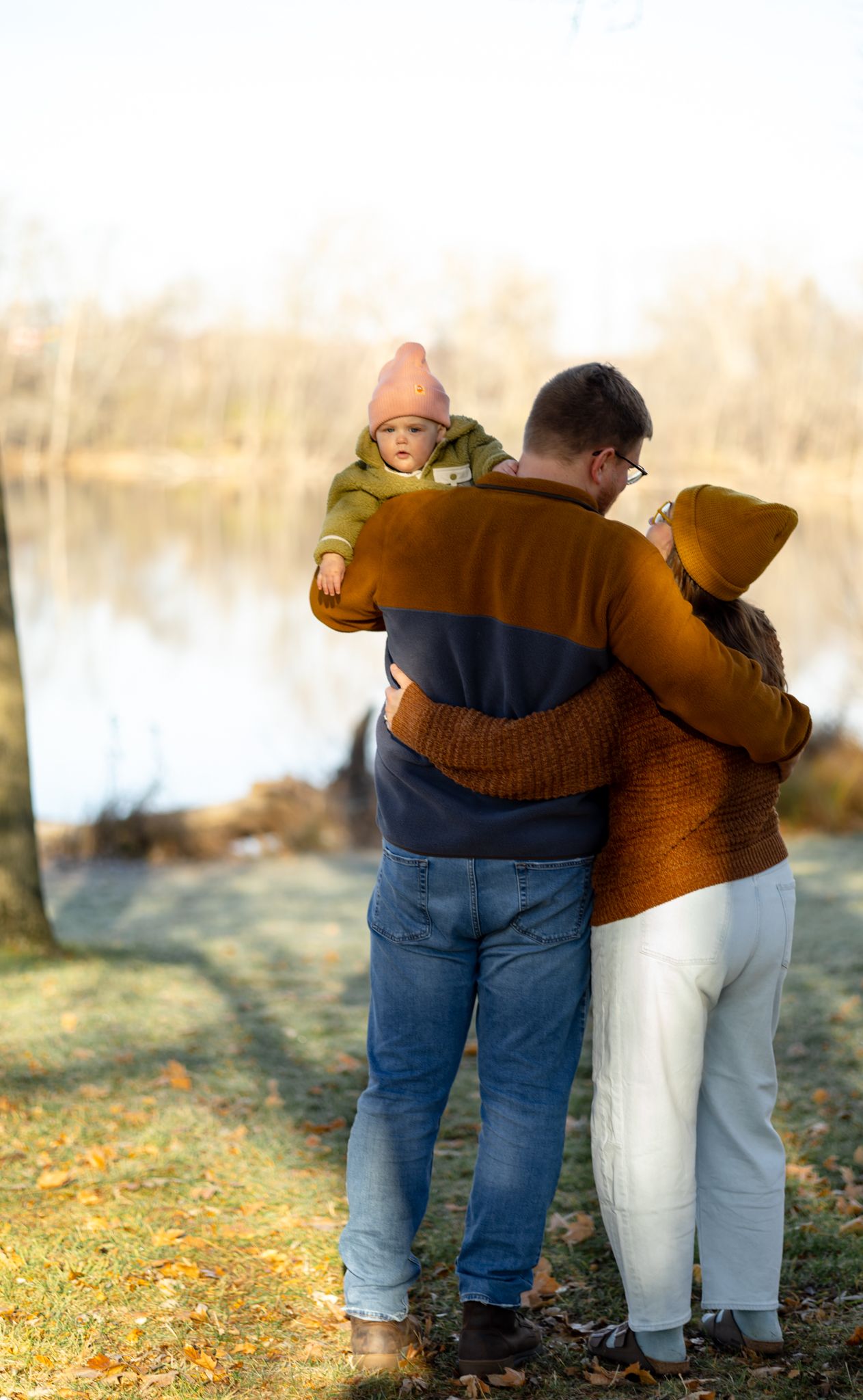 People in casual clothing walk together on fallen autumn leaves near a lake.