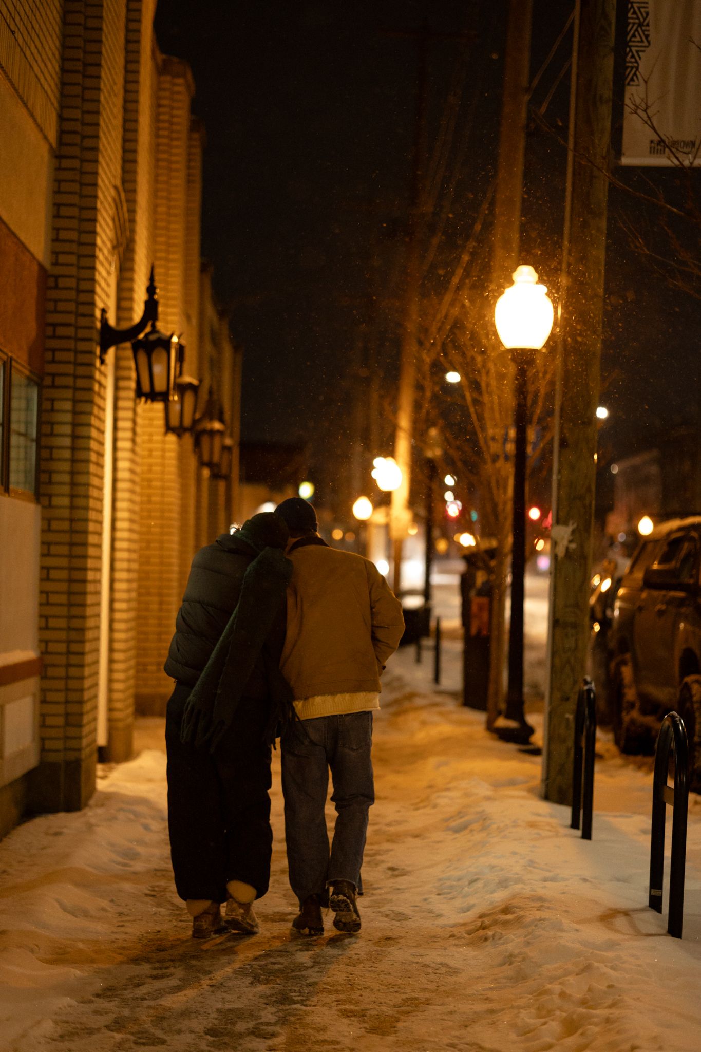 People walking along a snowy brick sidewalk lit by vintage streetlamps at night.