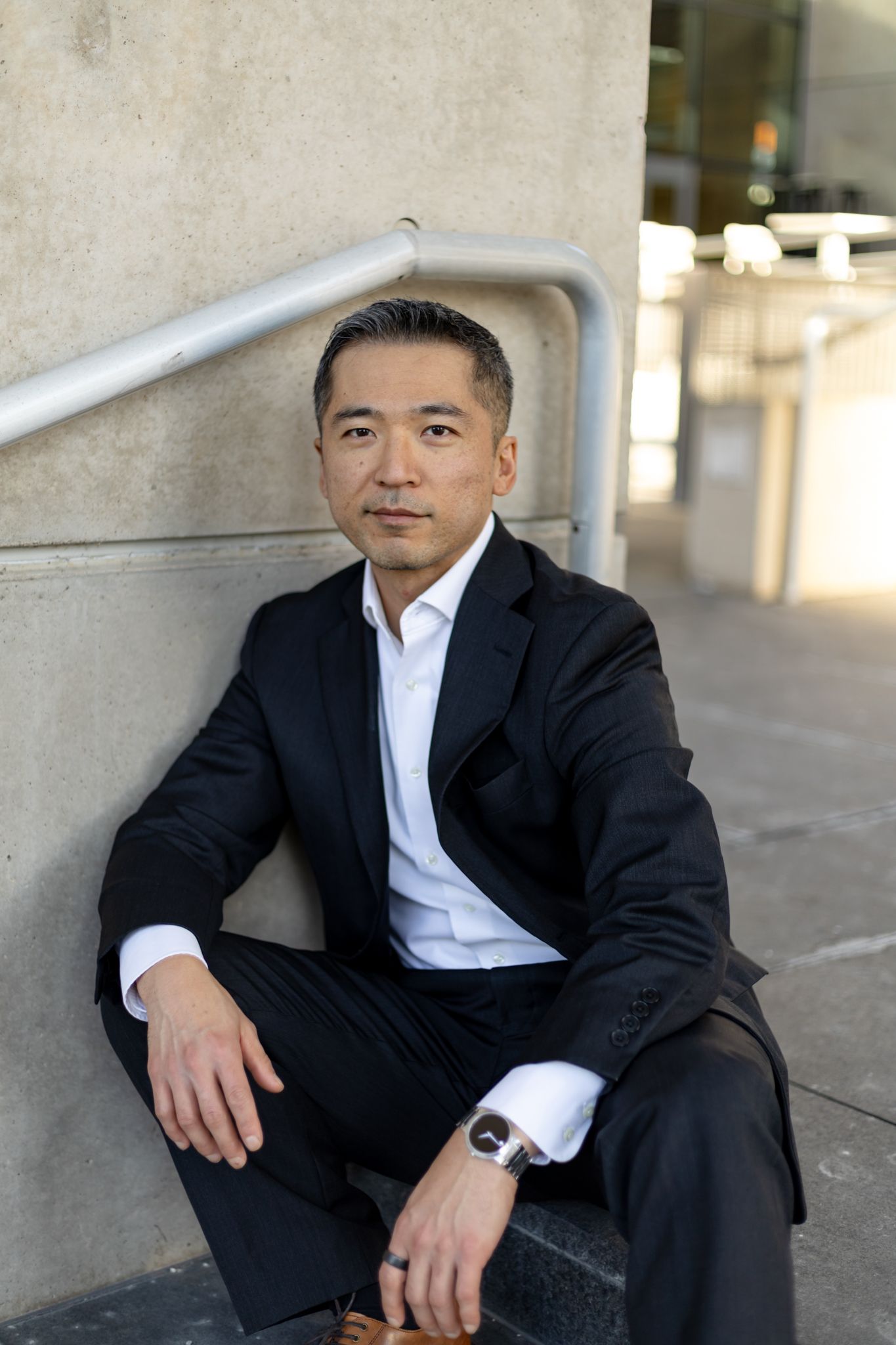 Professional in a black suit and white shirt sits casually against a concrete wall in an urban setting.