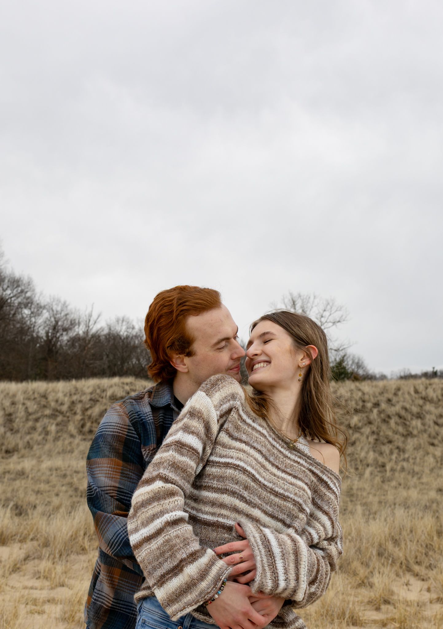 A couple embraces in a field, woman in striped sweater smiling as red-haired man leans in for a kiss.