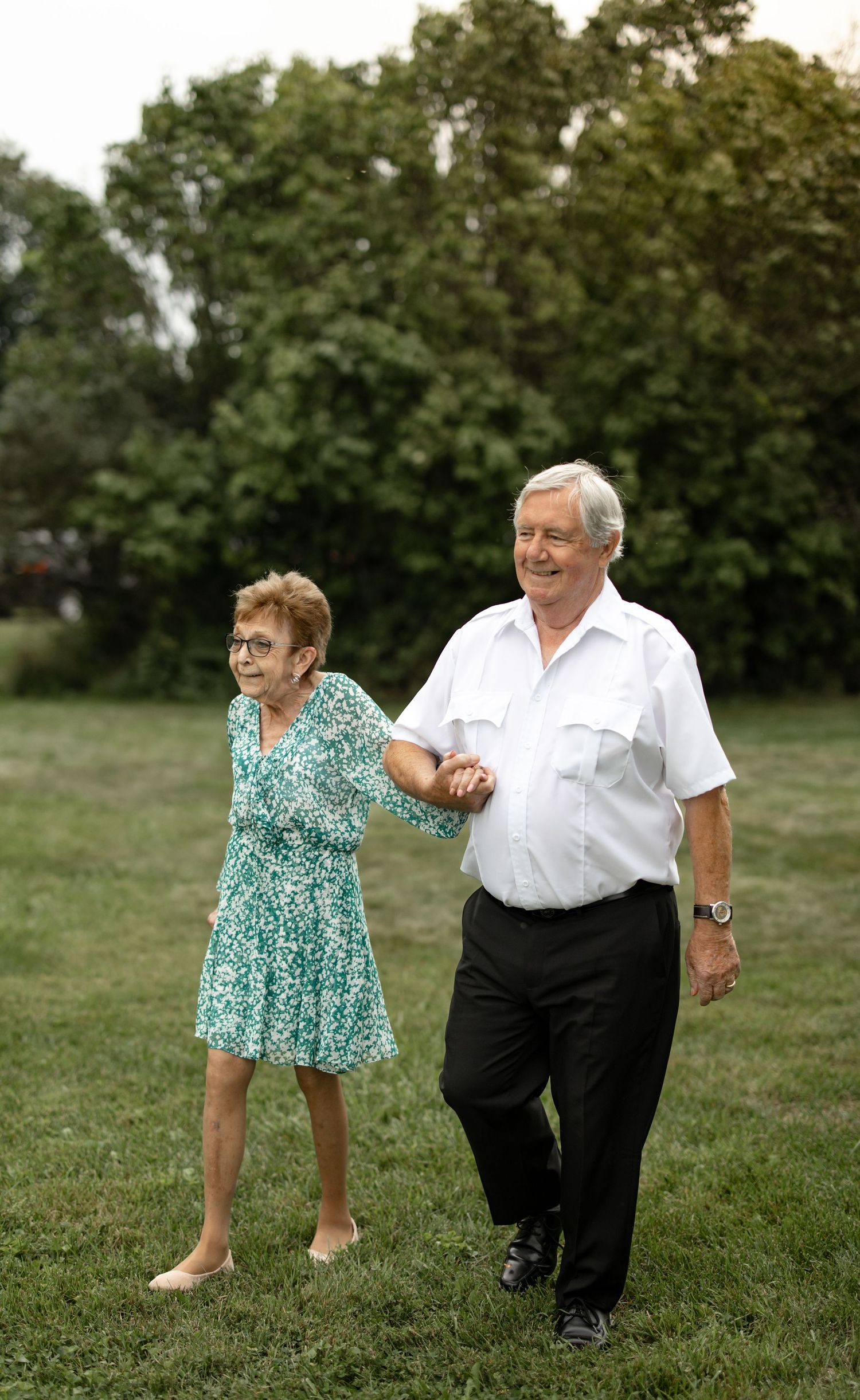 A couple dancing together on a grassy field surrounded by trees on a sunny day.