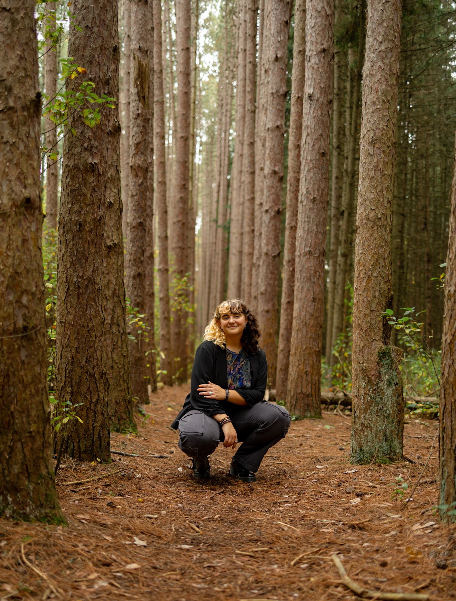 Figure crouching among tall pine trees in a dense forest setting.
