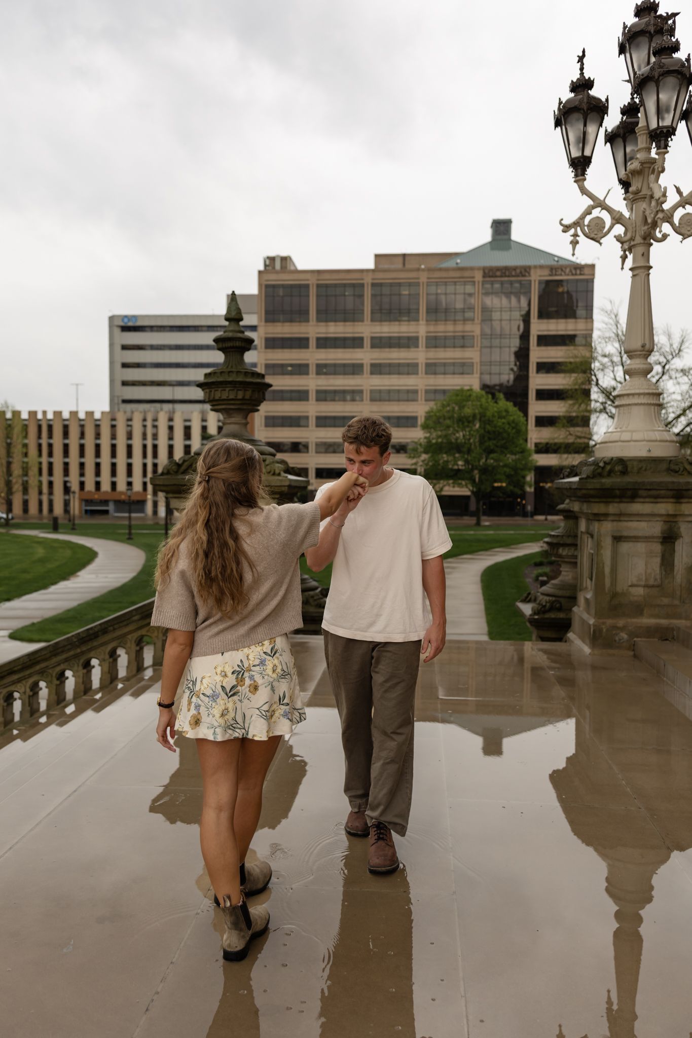 A couple walks on a wet terrace near a ornate lamp post, with city buildings in the background on a cloudy day.