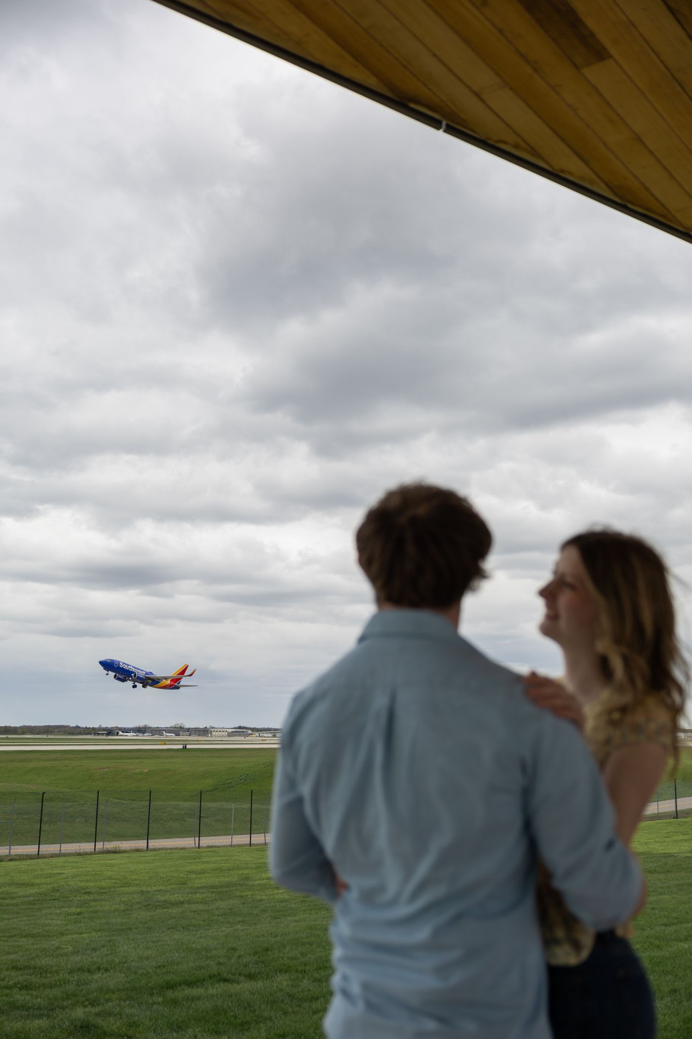 Couple watching a Southwest Airlines plane take off from an airport observation deck under cloudy skies.