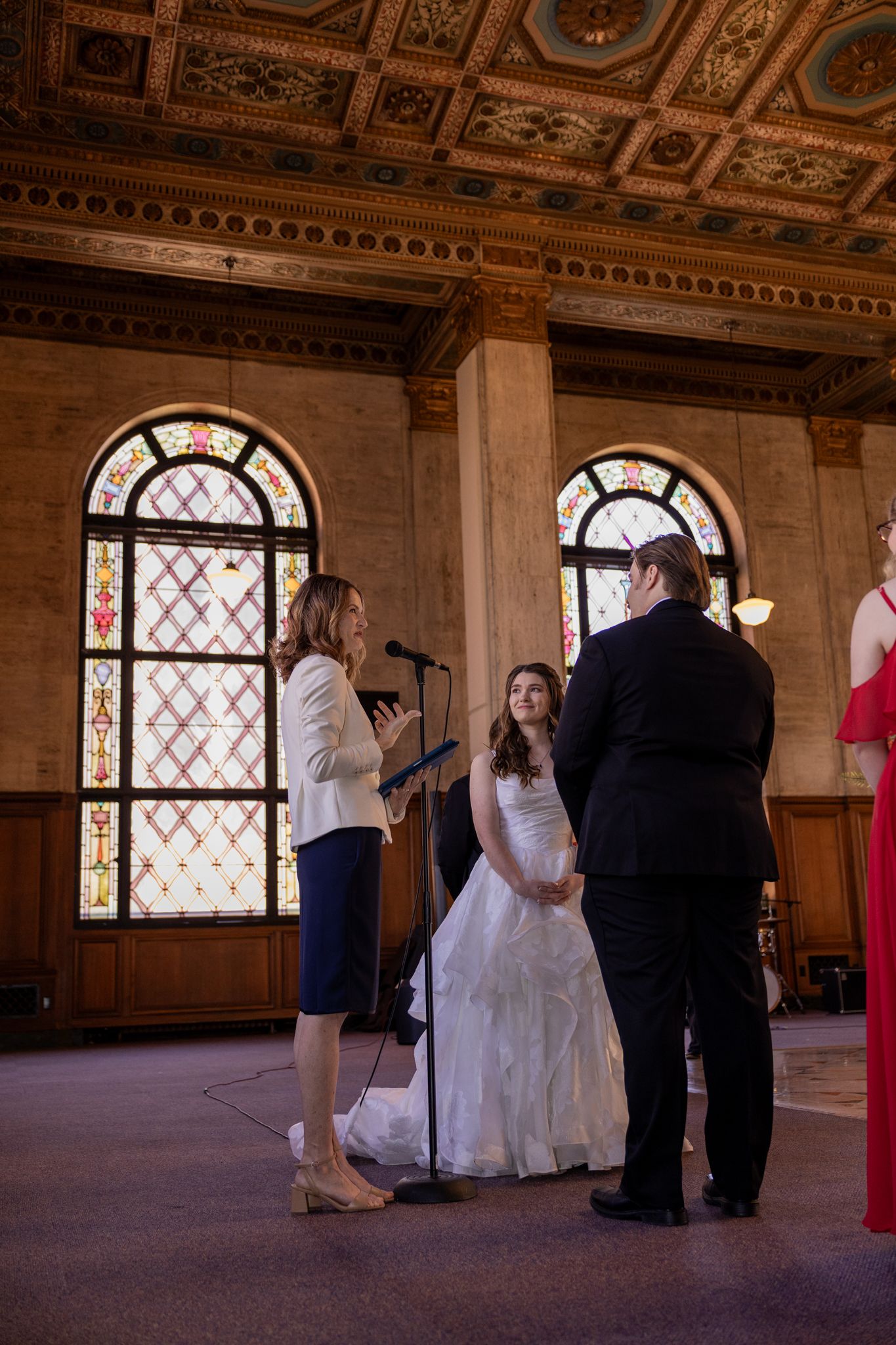 Wedding ceremony in ornate hall with stained glass windows, officiant reading vows to bride in white gown and groom in black suit.