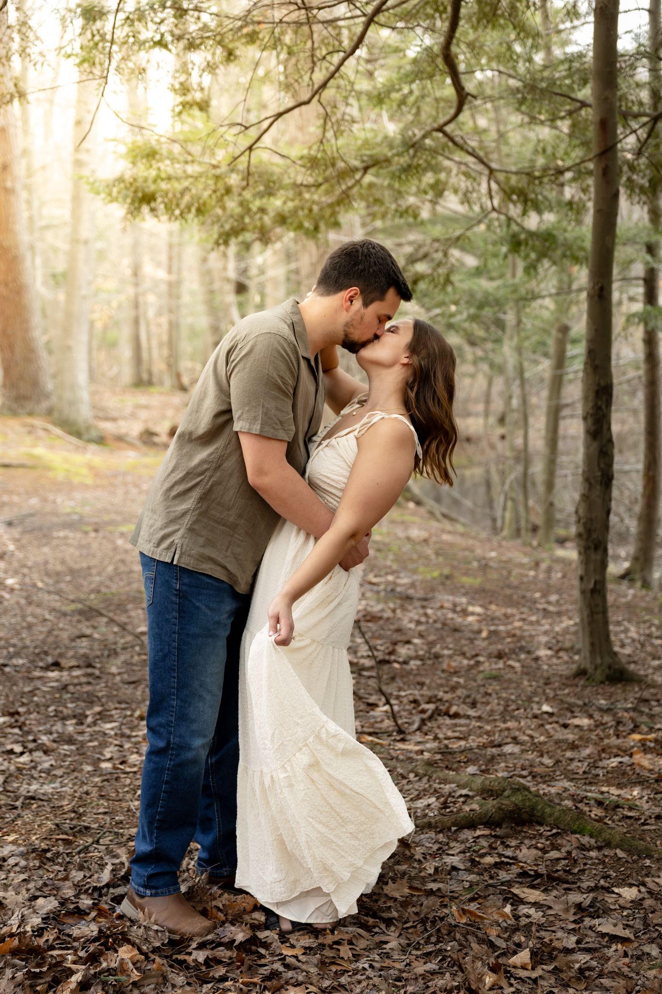A couple shares a romantic kiss in a sun-dappled forest, the woman in a white dress and man in a green shirt and jeans.
