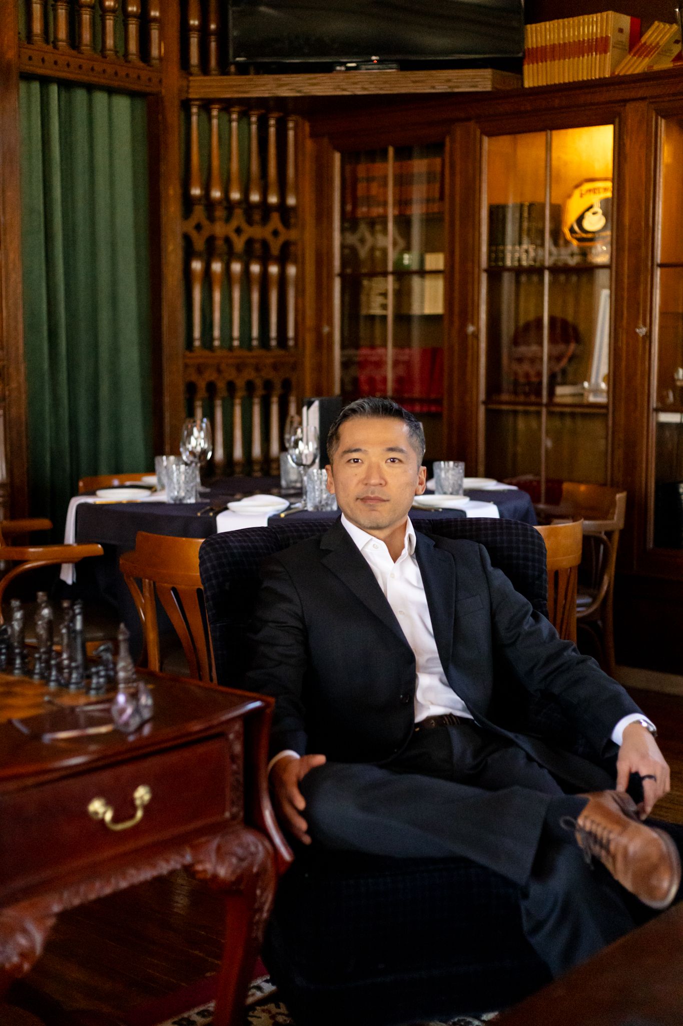 A person in a black suit relaxes on a leather couch in an elegant wood-paneled room with warm lighting.
