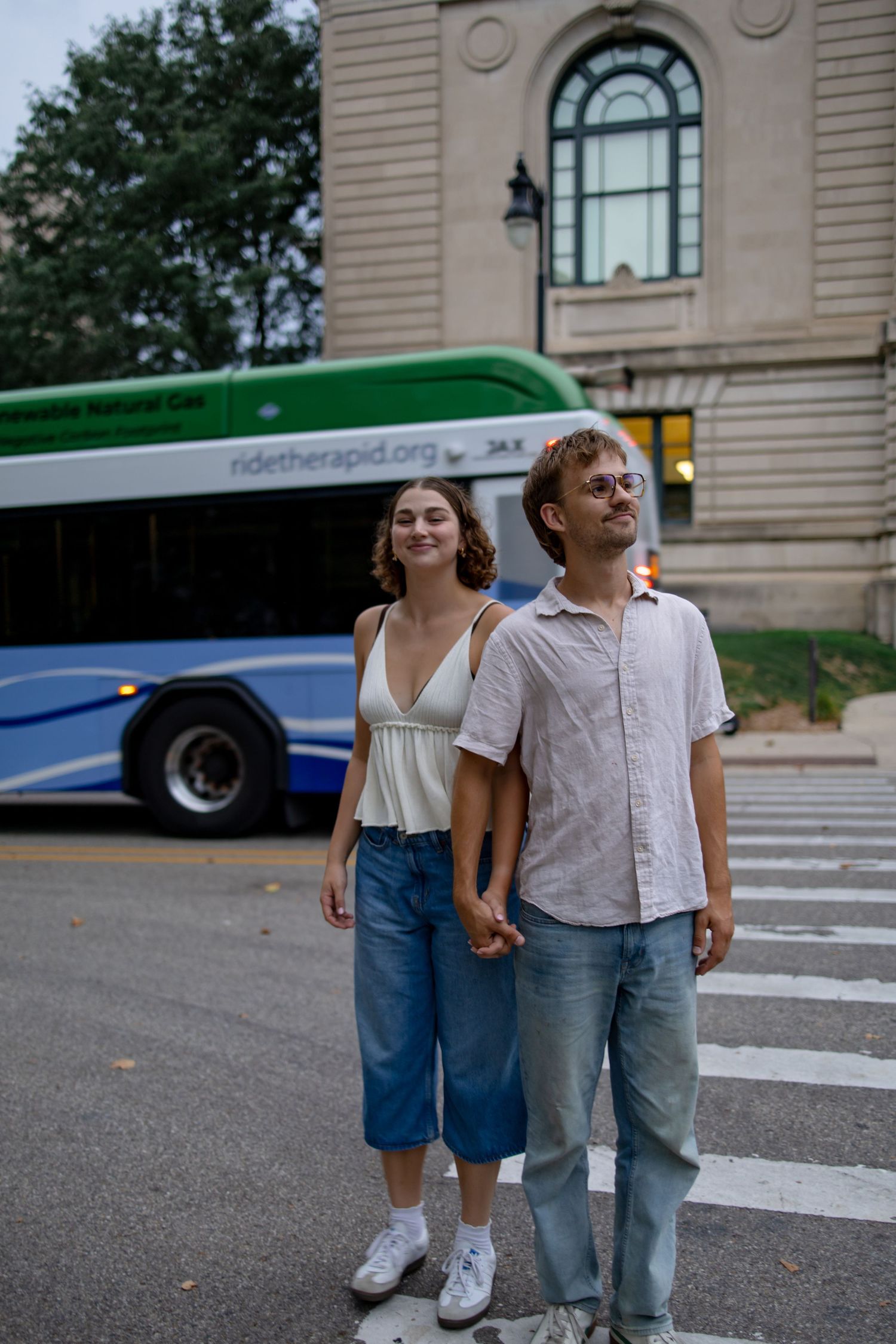 A couple poses casually in front of a historic building with buses visible in the background.