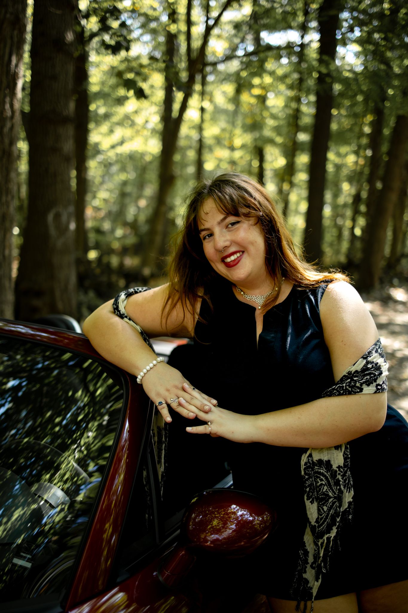 Playful poses with red convertible car in sunlit forest setting.