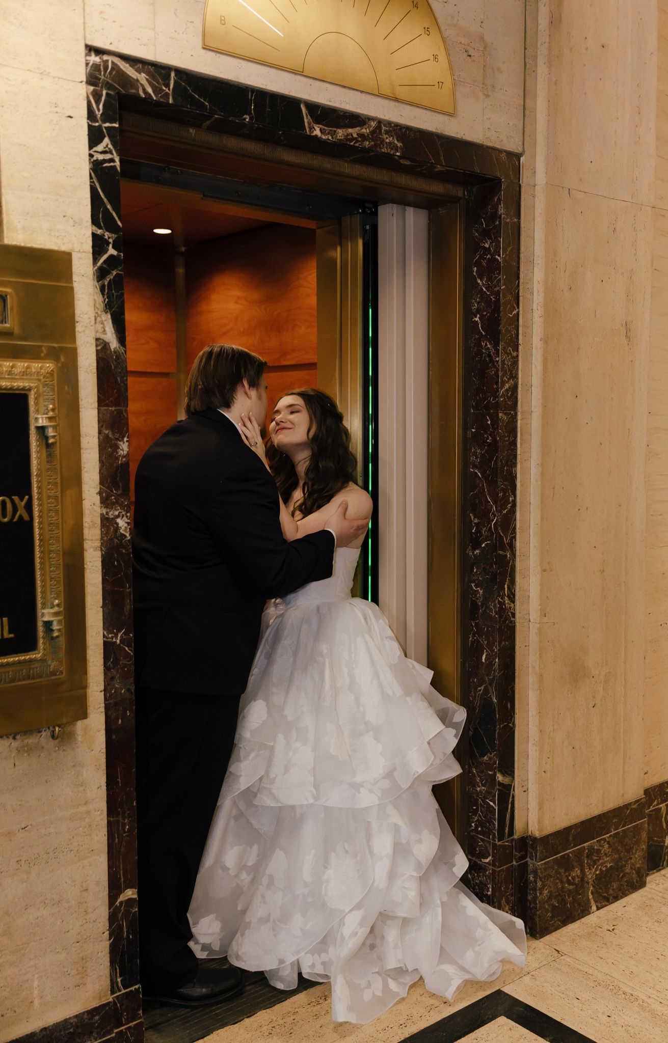 Bride in layered white gown and groom in black tuxedo share a romantic moment in an elegant elevator doorway.