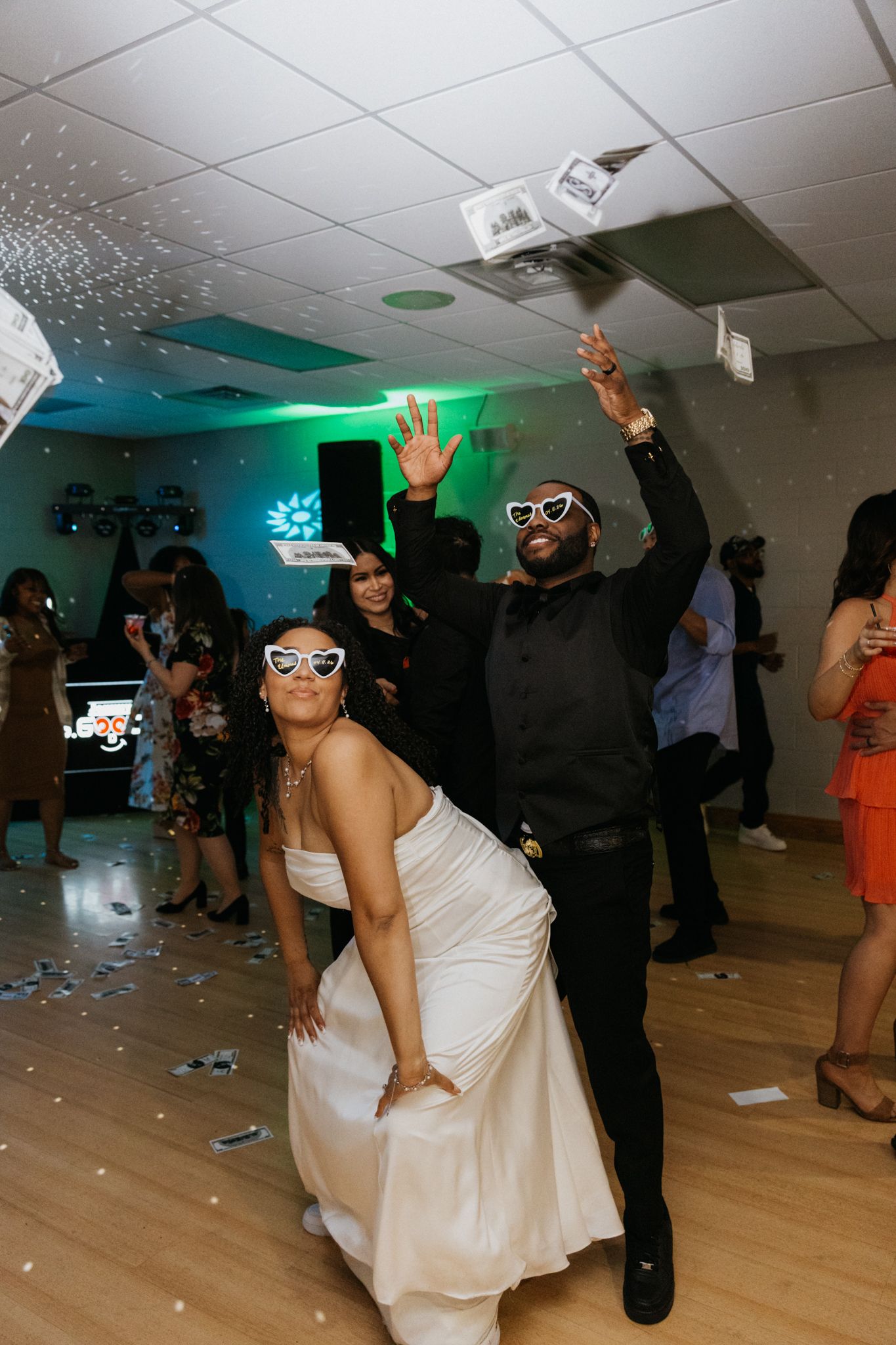 Bride in white gown and groom in black suit tossing money on dance floor at wedding reception.