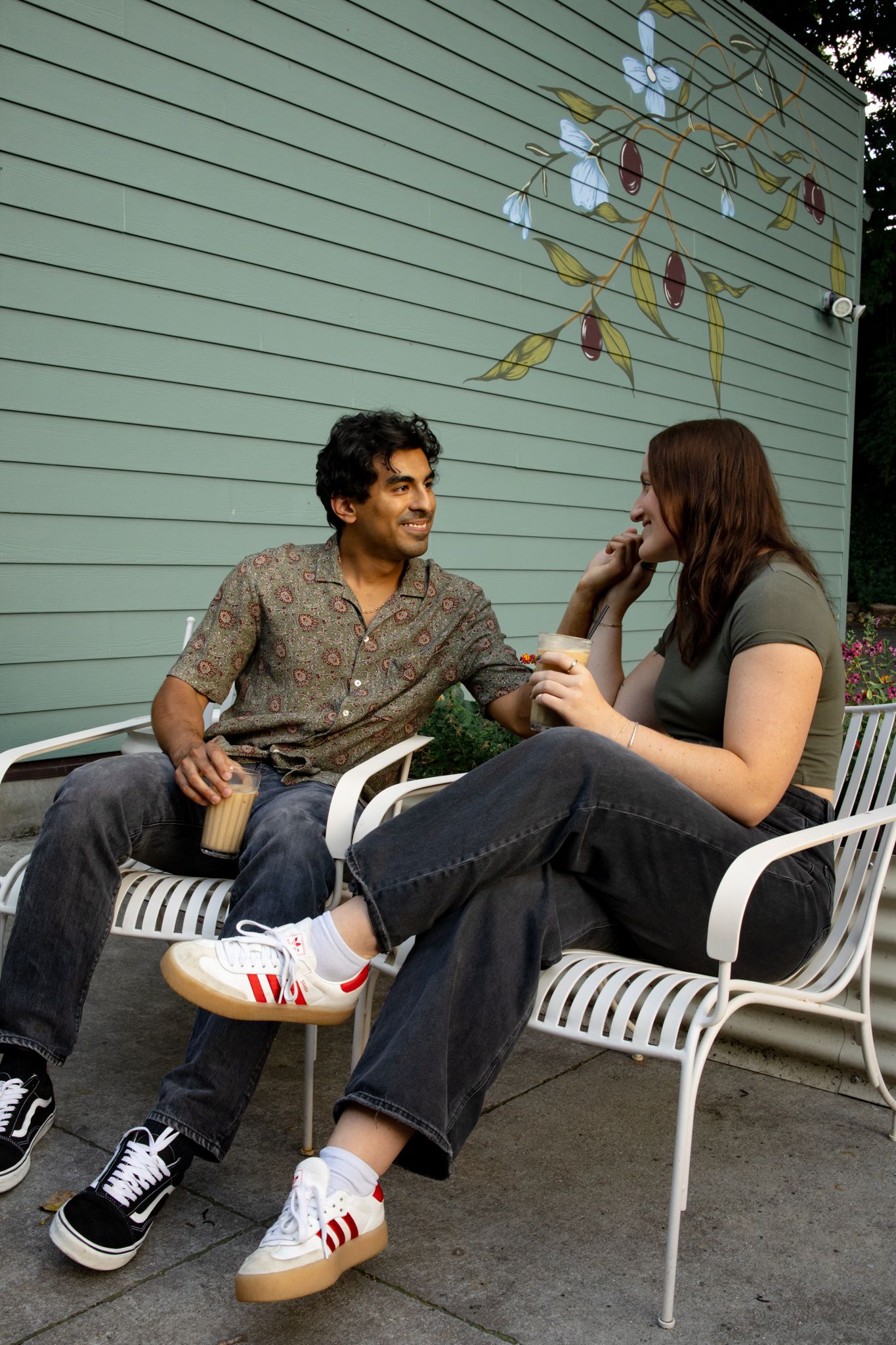 Two people chat while relaxing on white metal chairs against painted wall outdoors.