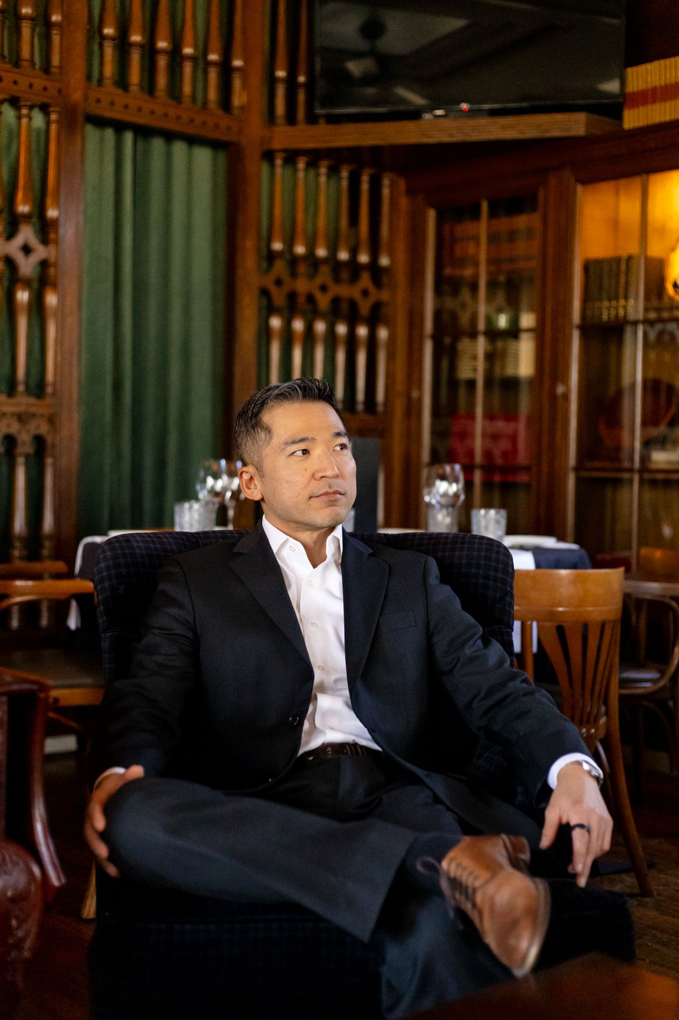 A person in a dark business suit sits confidently in a leather chair at an upscale bar or restaurant with wood paneling.