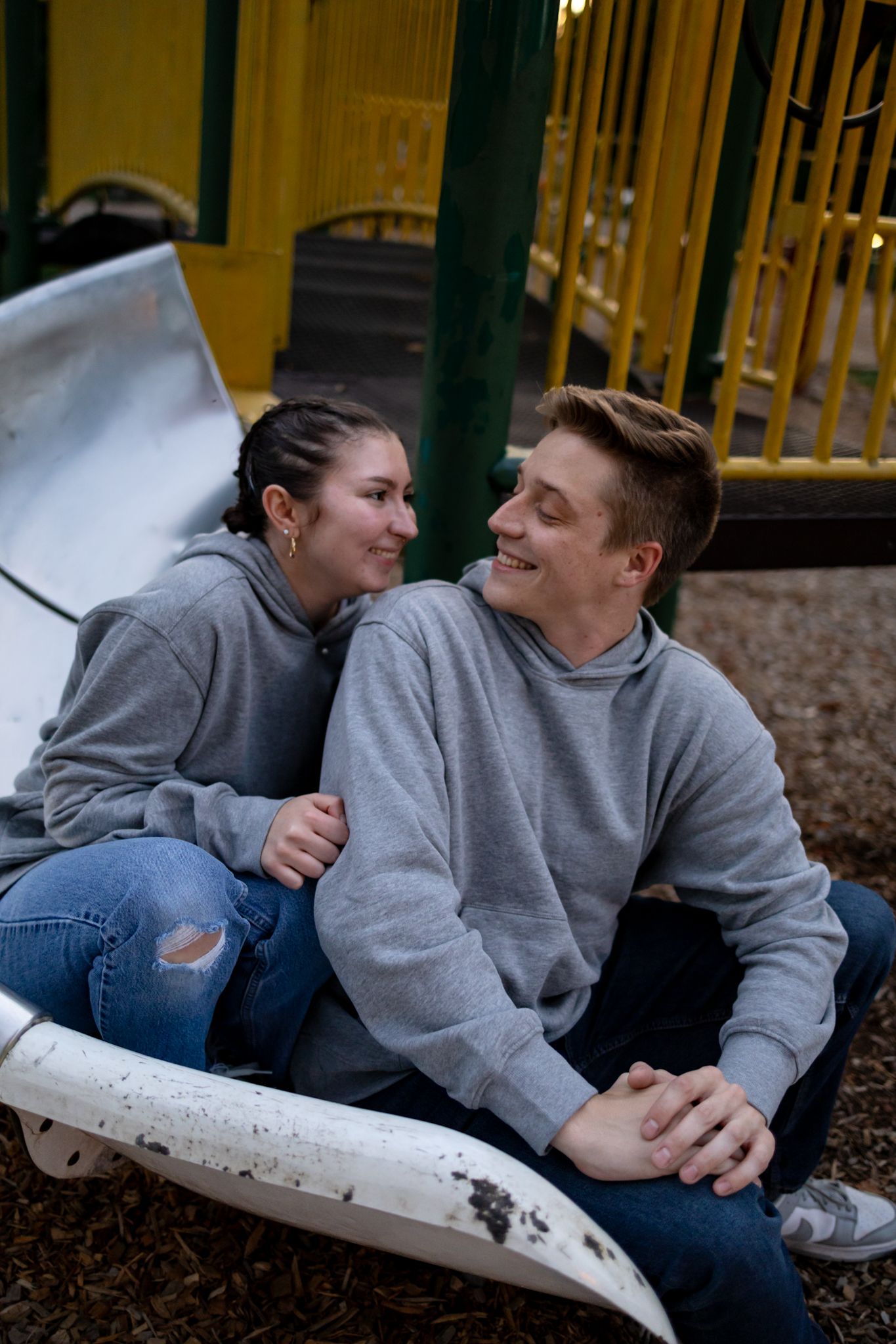 Two people in matching gray hoodies share a playful moment on a playground slide.