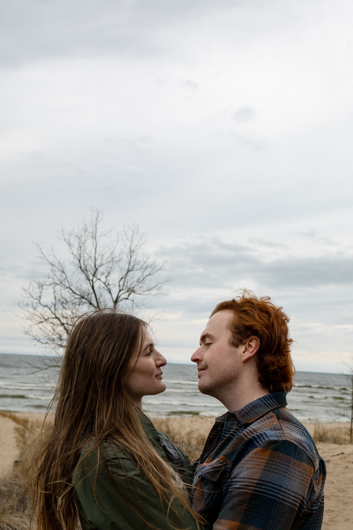A couple smiles at each other on a sandy beach with a bare tree and cloudy sky behind them.