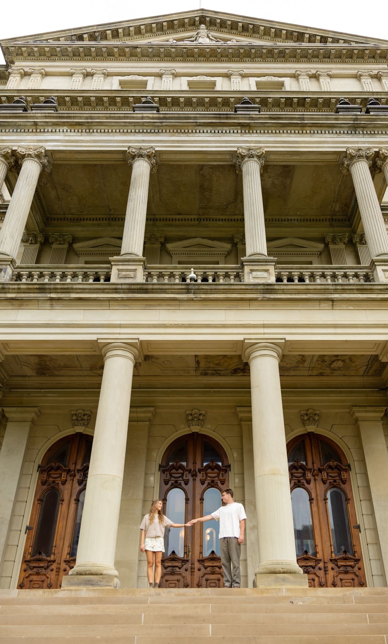 Couple holding hands on steps of grand neoclassical building with tall columns and ornate wooden doors.
