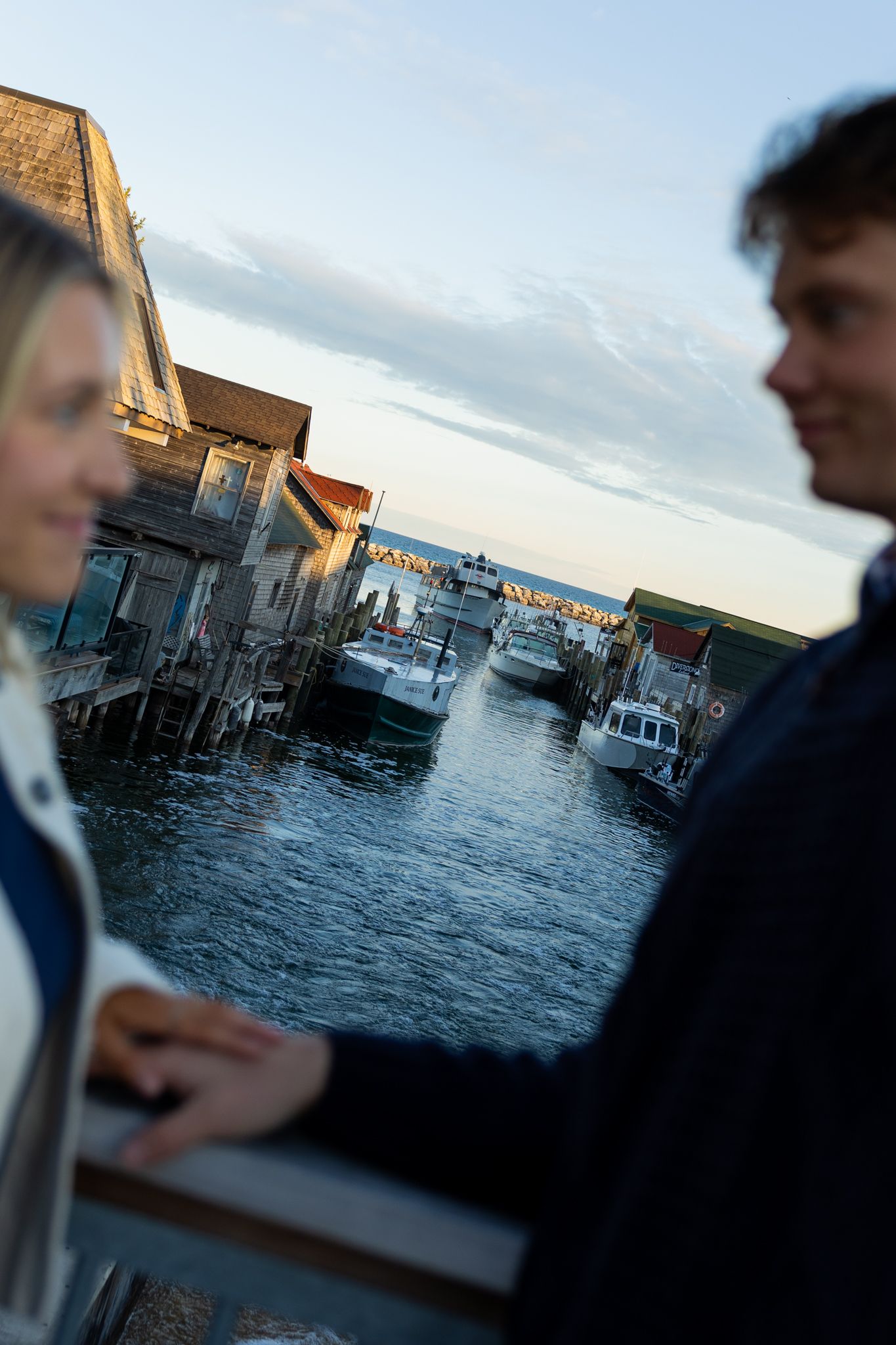 Couple holding hands on a waterfront dock at sunset with boats and wooden buildings reflected in calm harbor waters.