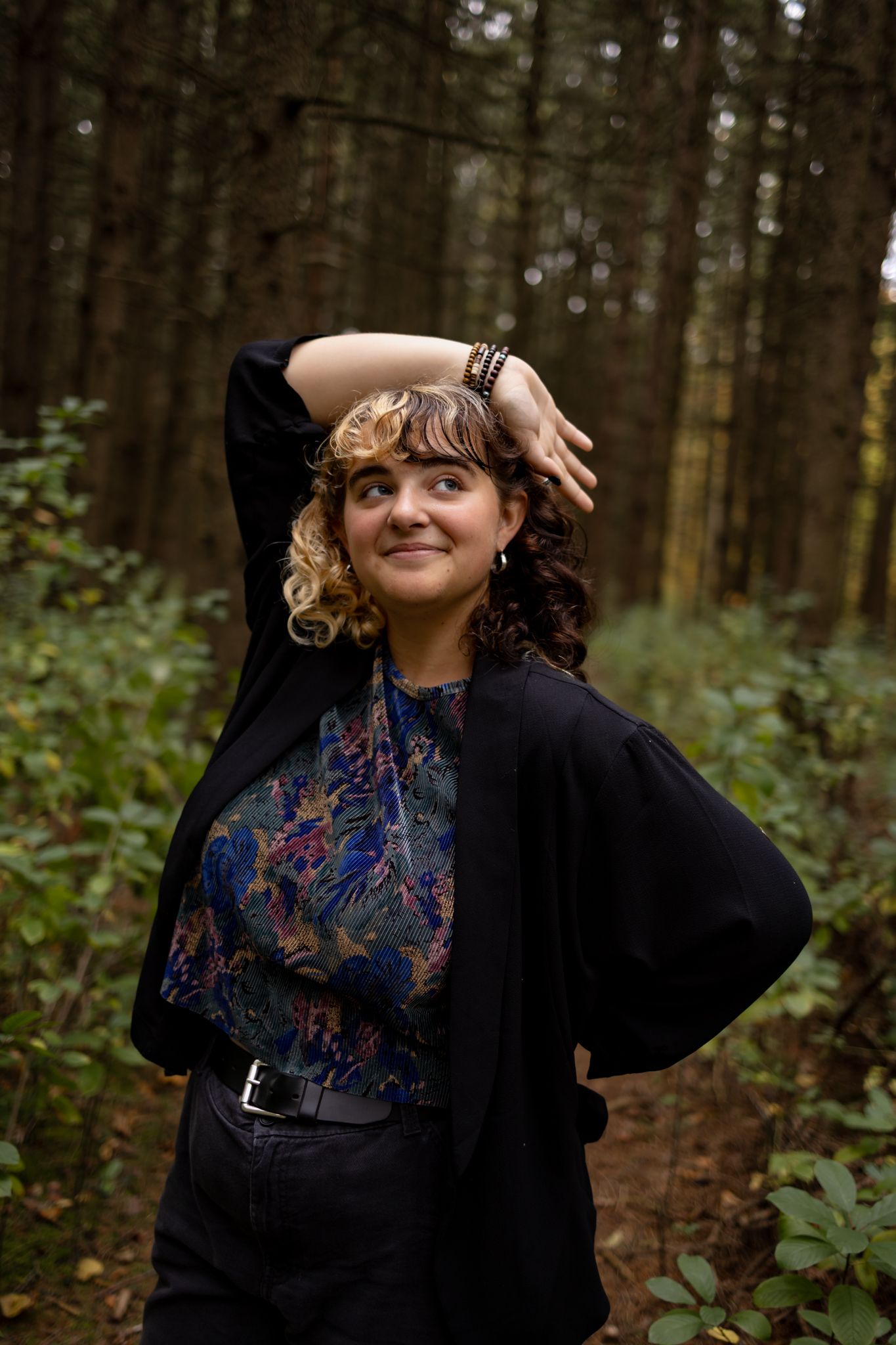 Person in black clothing poses playfully against a forest backdrop.