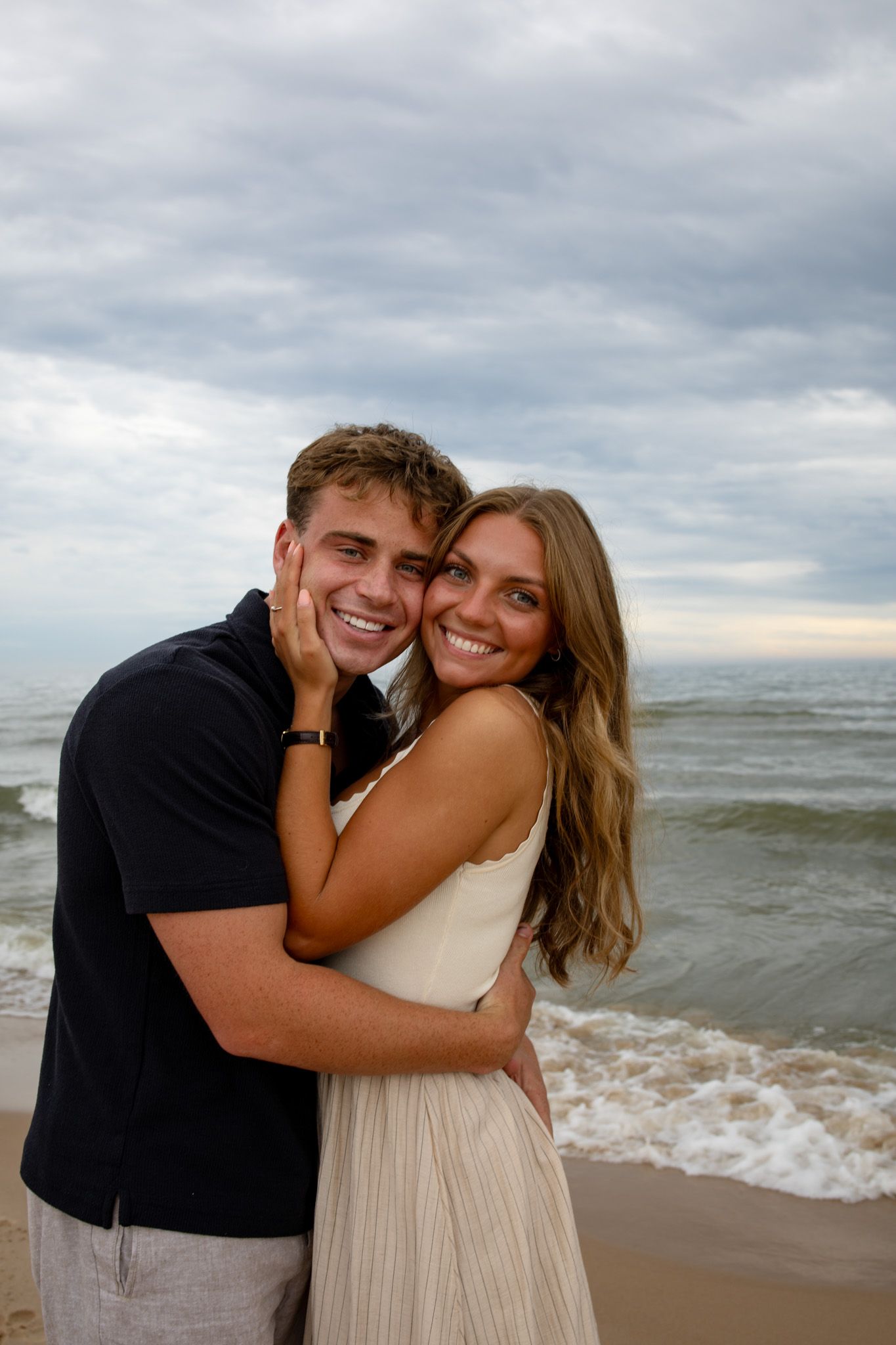 A joyful couple embraces on a cloudy beach at sunset.