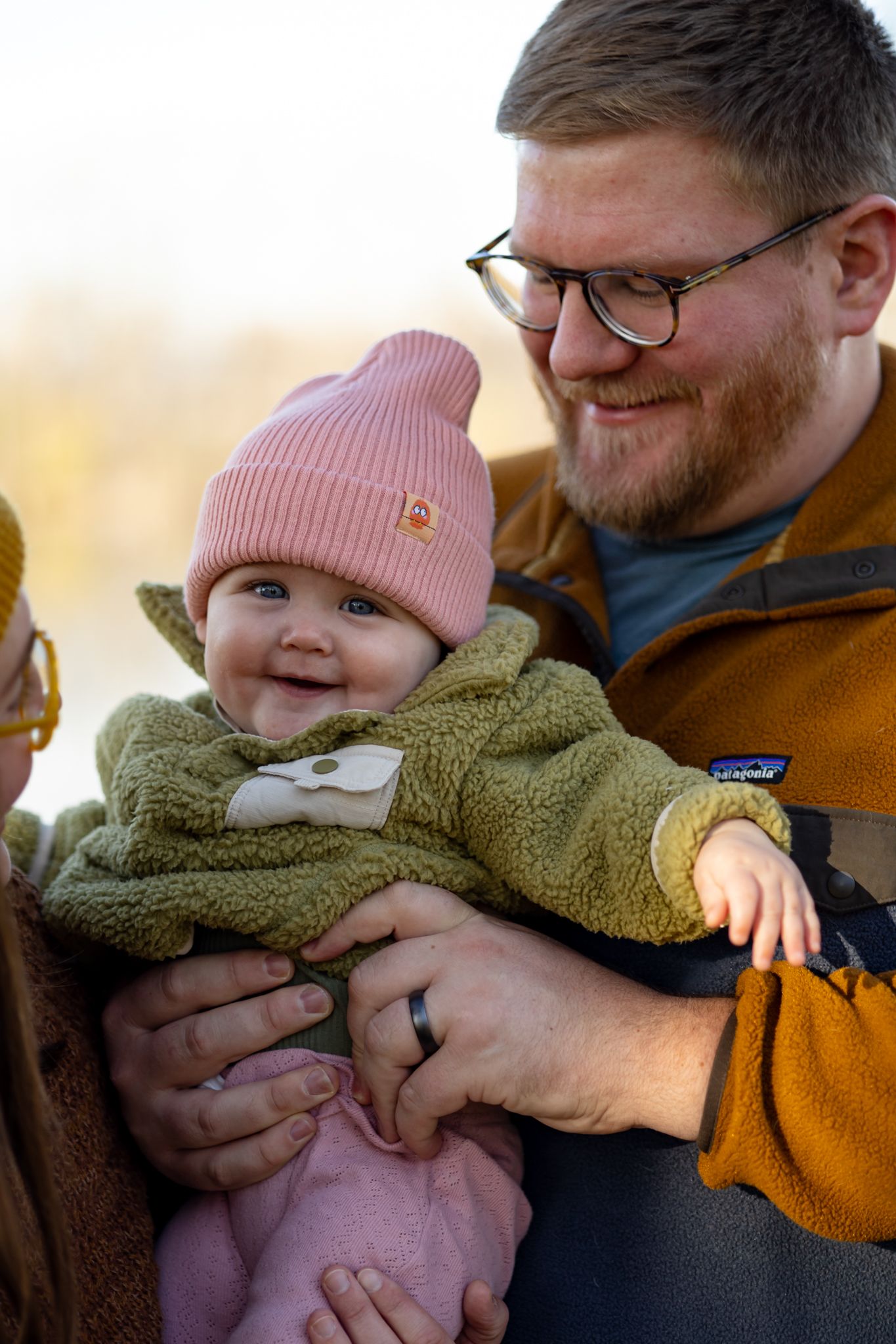 A tender family moment captured outdoors with a baby wearing a pink hat and olive green fleece.