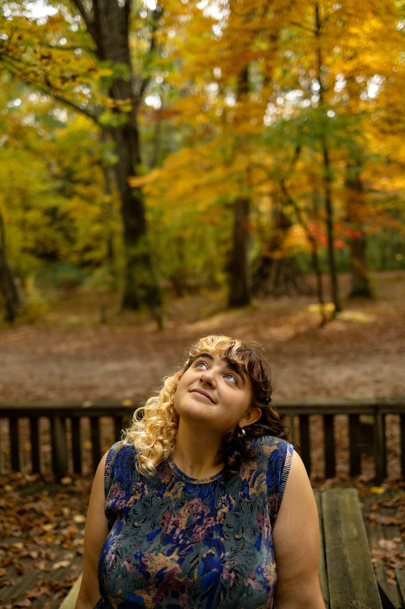 A person in a floral dress sits on a bench surrounded by autumn foliage.