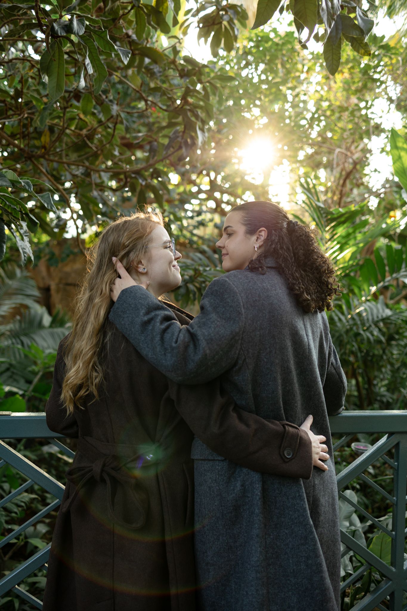 Two people in dark coats share an intimate moment in a garden with sunlight streaming through the trees.