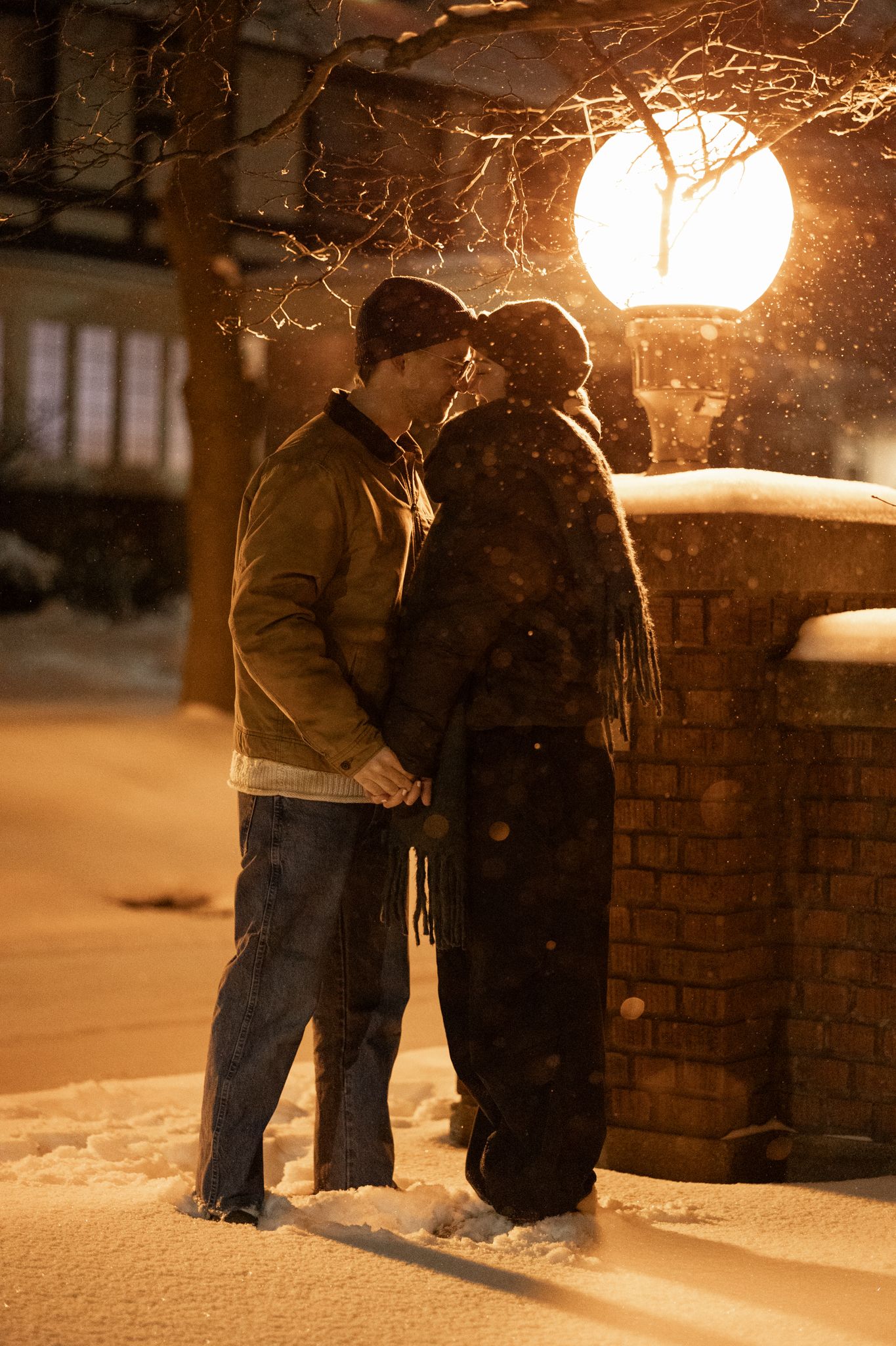 Couple standing near a brick wall in the snow under warm streetlight.