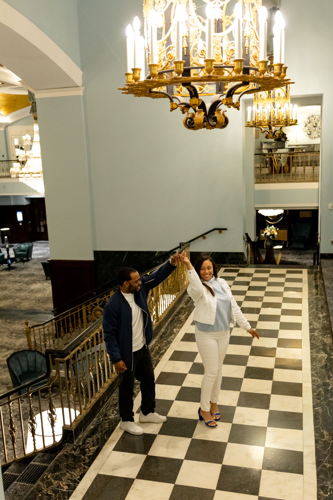 Couple posing under ornate gold chandelier in elegant hotel lobby with black and white checkered marble floors.