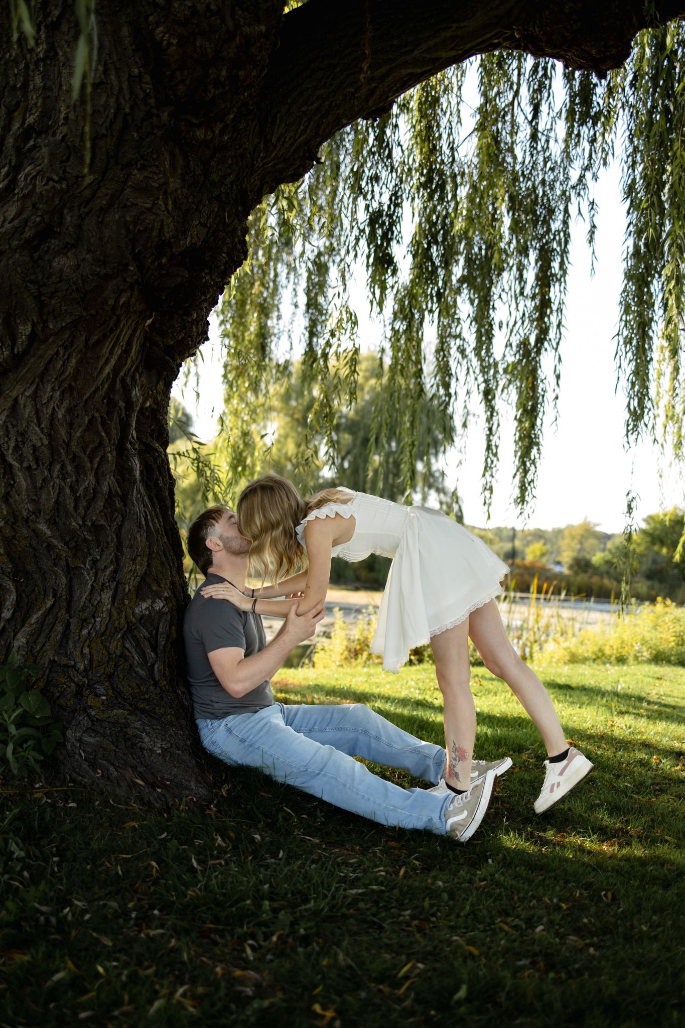 A couple reclines together under a weeping willow tree during a summer photoshoot.