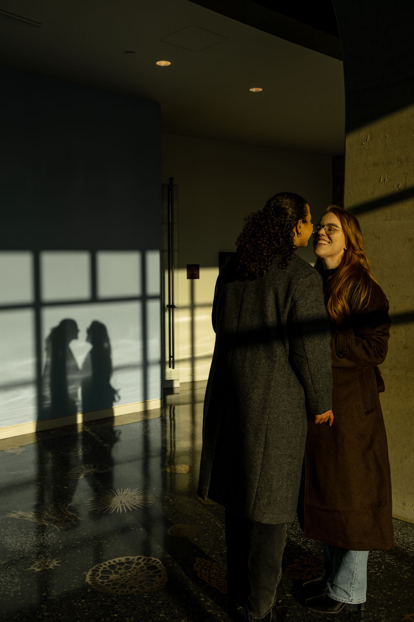 Two people in long coats embrace in a dimly lit hallway while their shadows cast on the wall behind them.