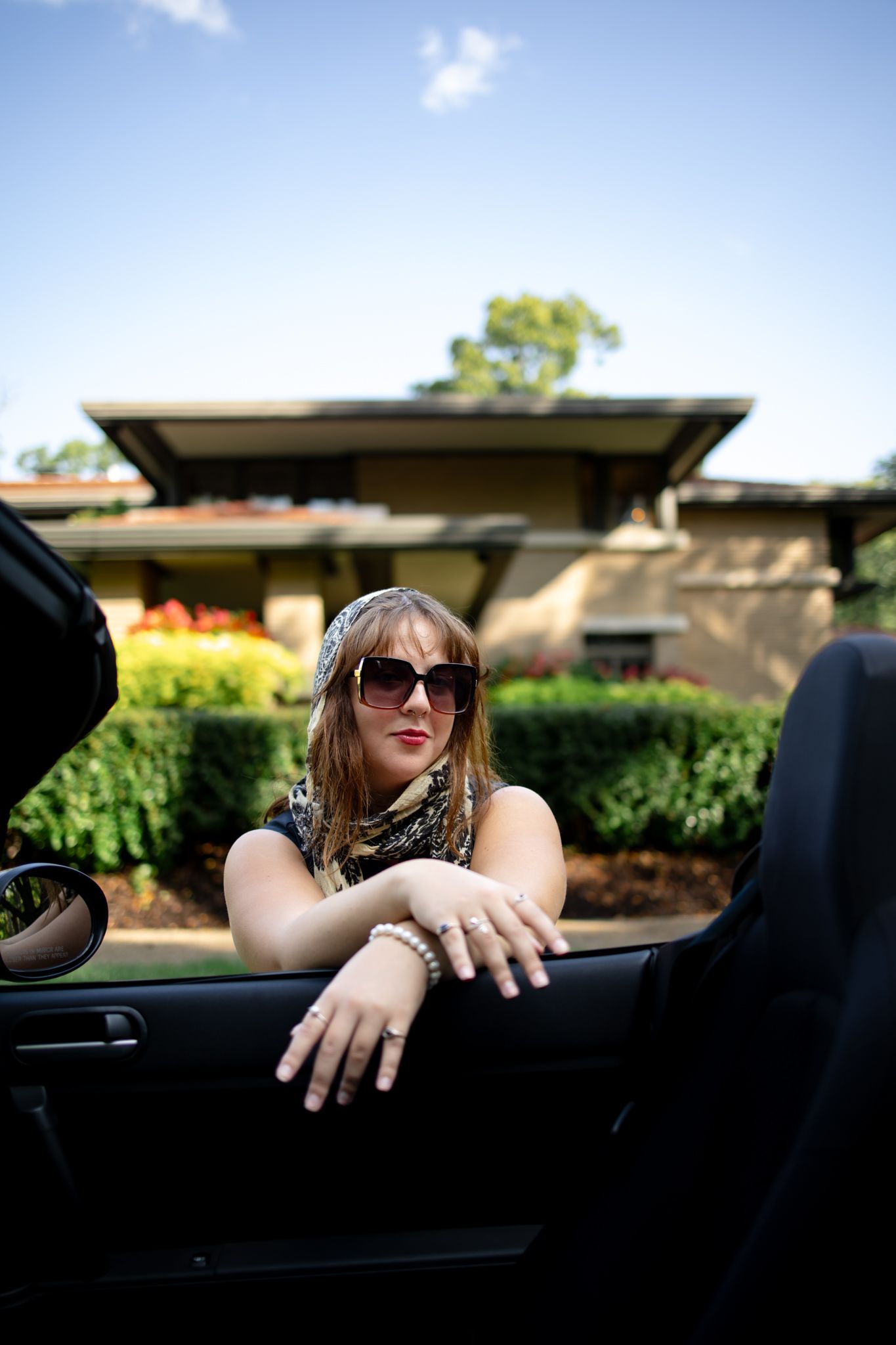 Someone wearing sunglasses relaxes in a convertible car on a sunny day with trees in the background.