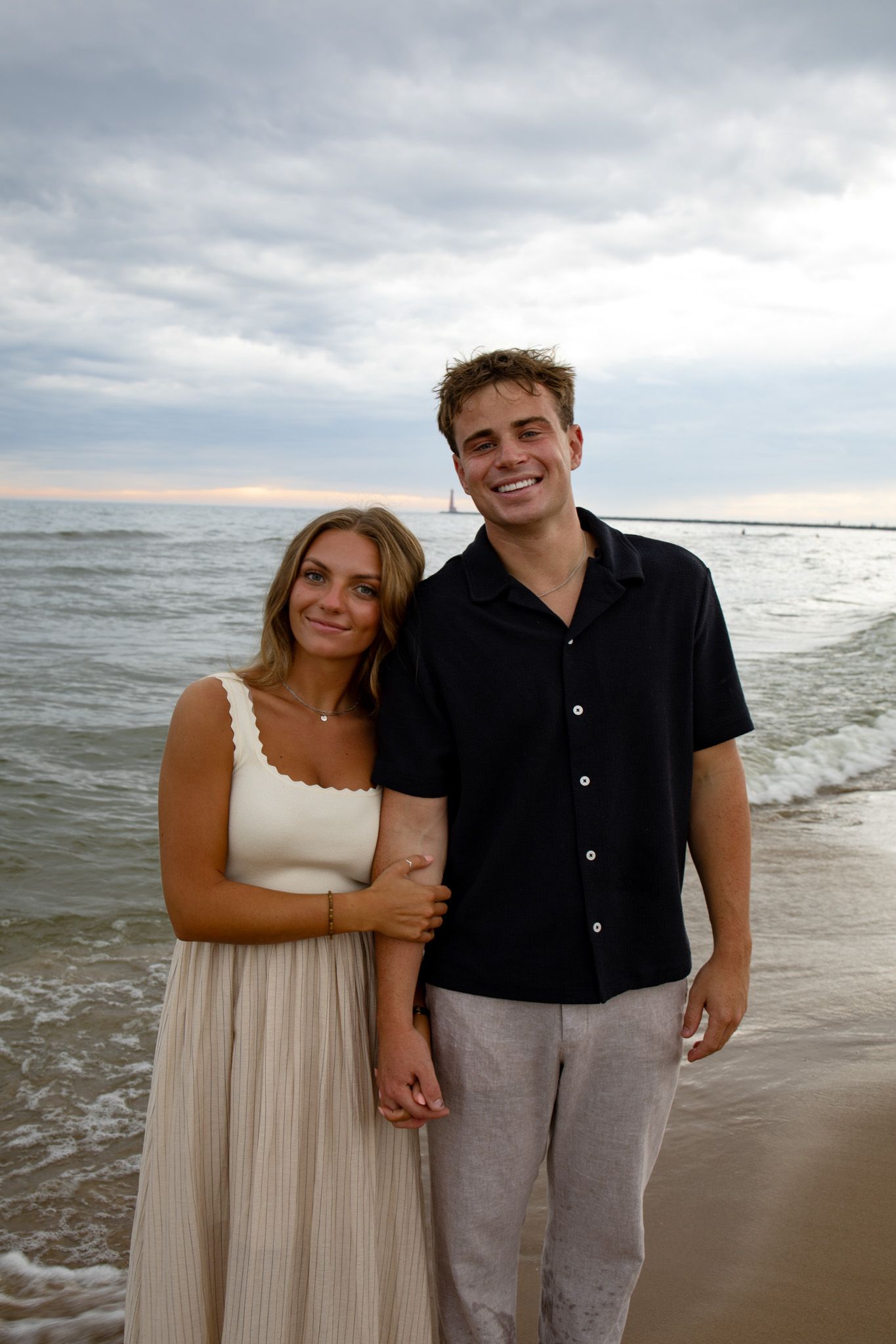 A couple poses together on a beach with waves and cloudy skies in the background.