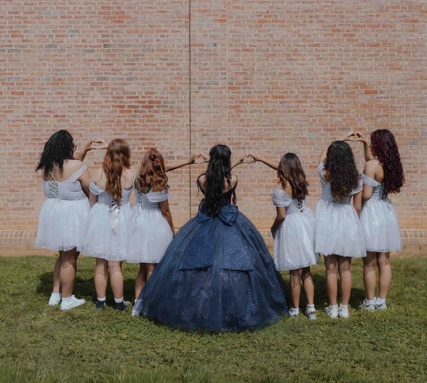 Group of girls in white dresses forming a circle with their hair braided together near a brick wall.