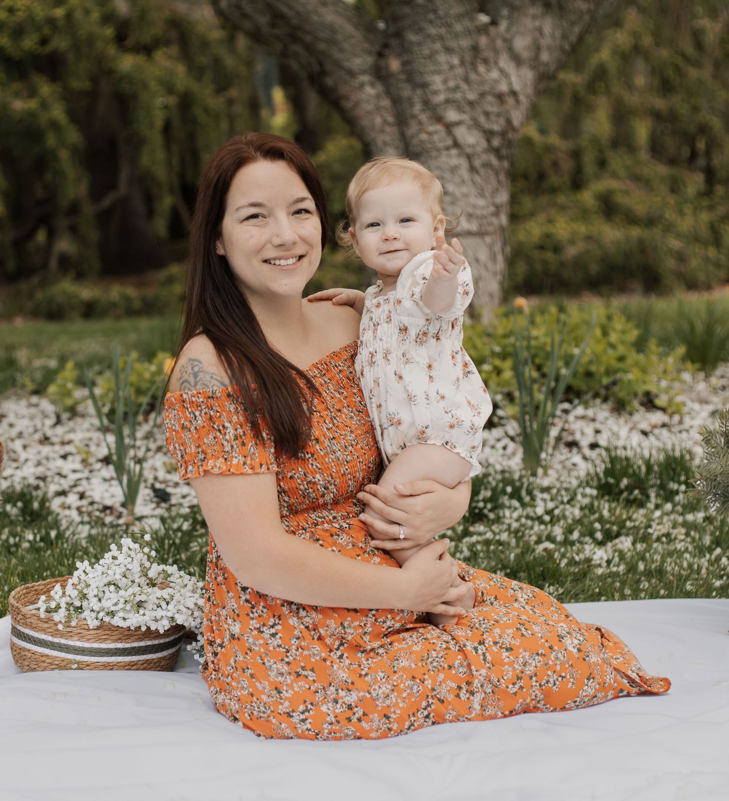 Smiling mother in orange floral dress sitting outdoors holding toddler on white blanket.