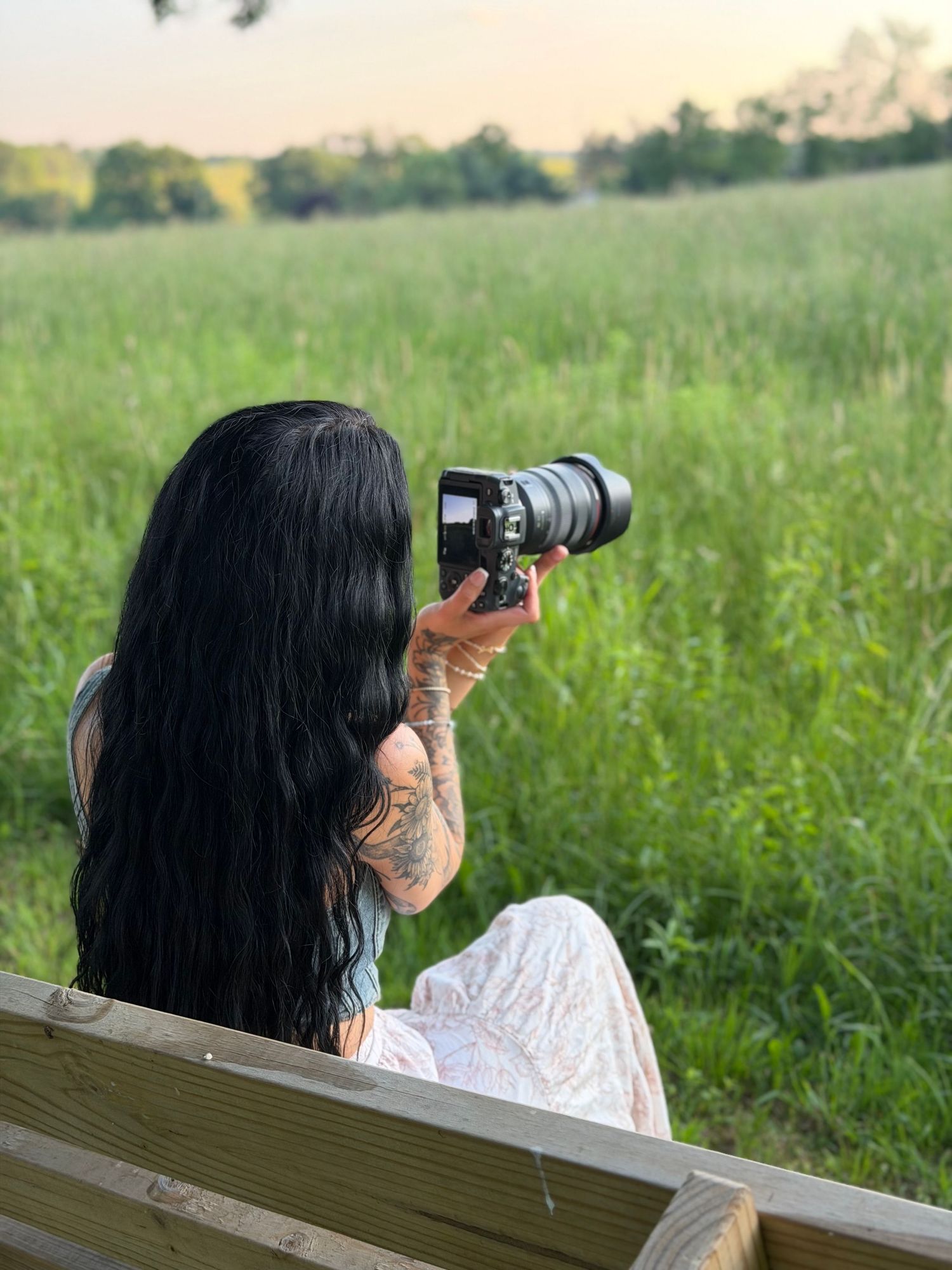 Tattooed woman with long black hair sitting on bench photographing green field at sunset with DSLR camera.