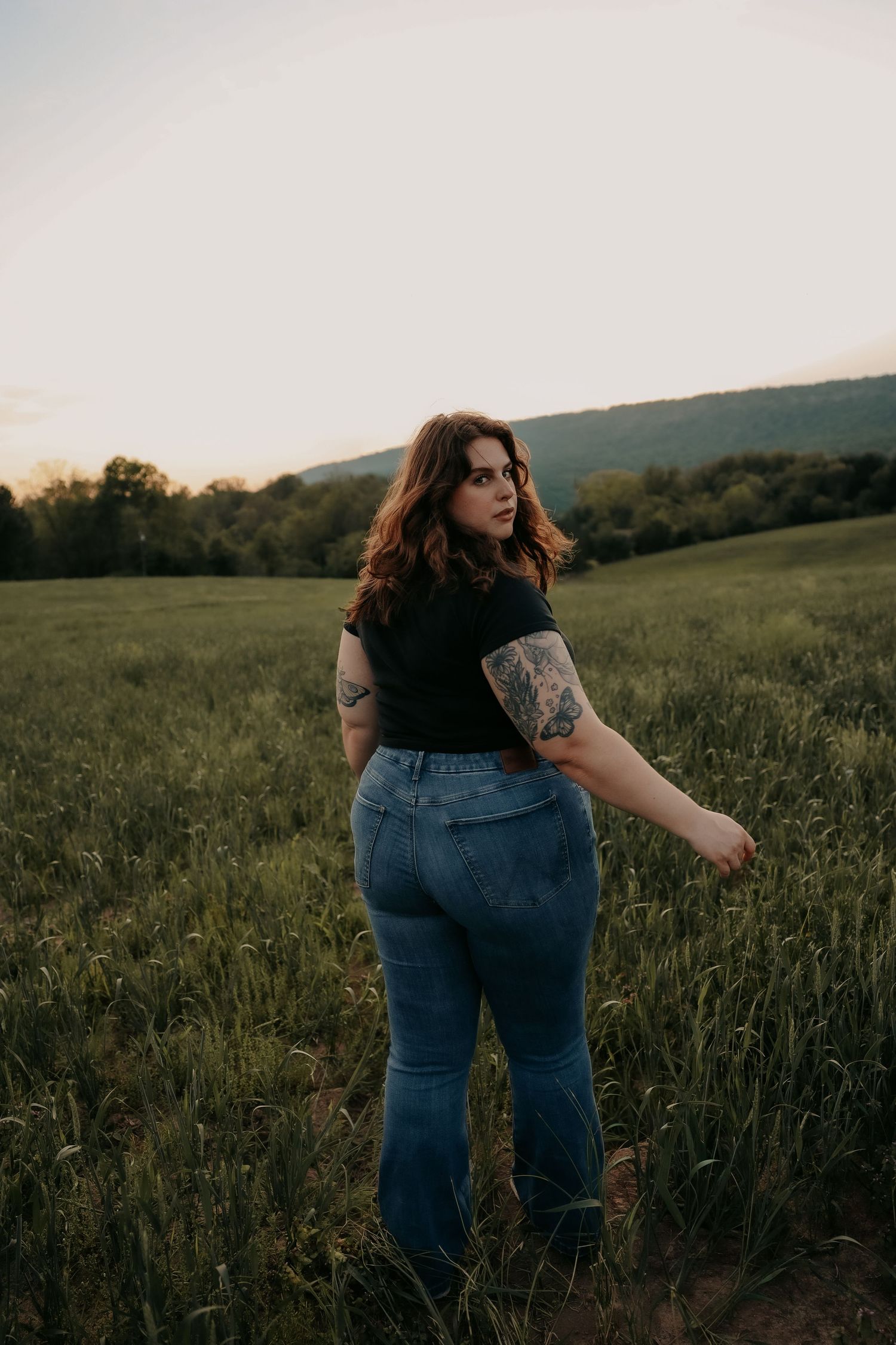 Woman with tattoos in black top and jeans standing in green field at sunset with mountain backdrop.