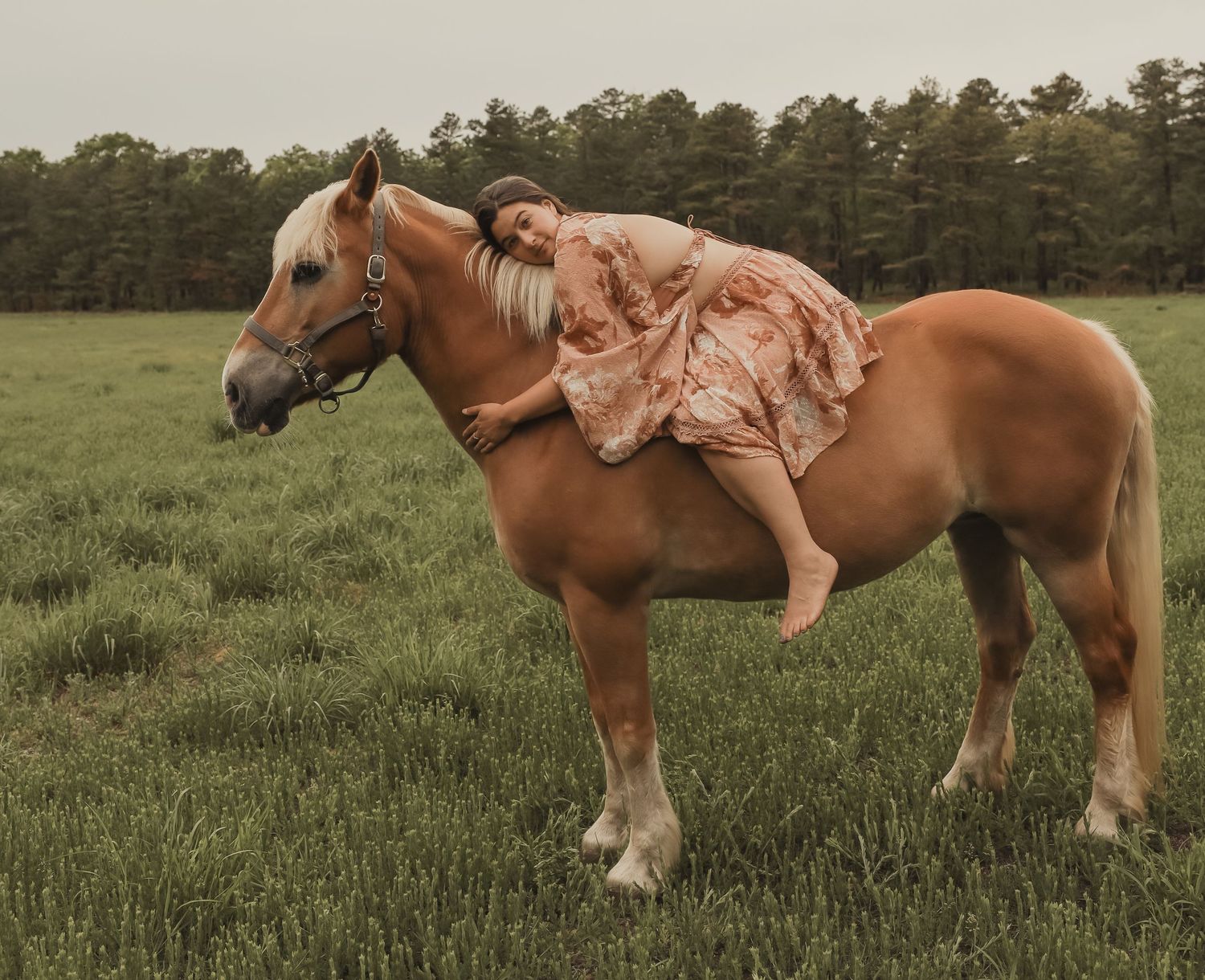 Woman in pink floral dress leaning forward on palomino horse in open green field.
