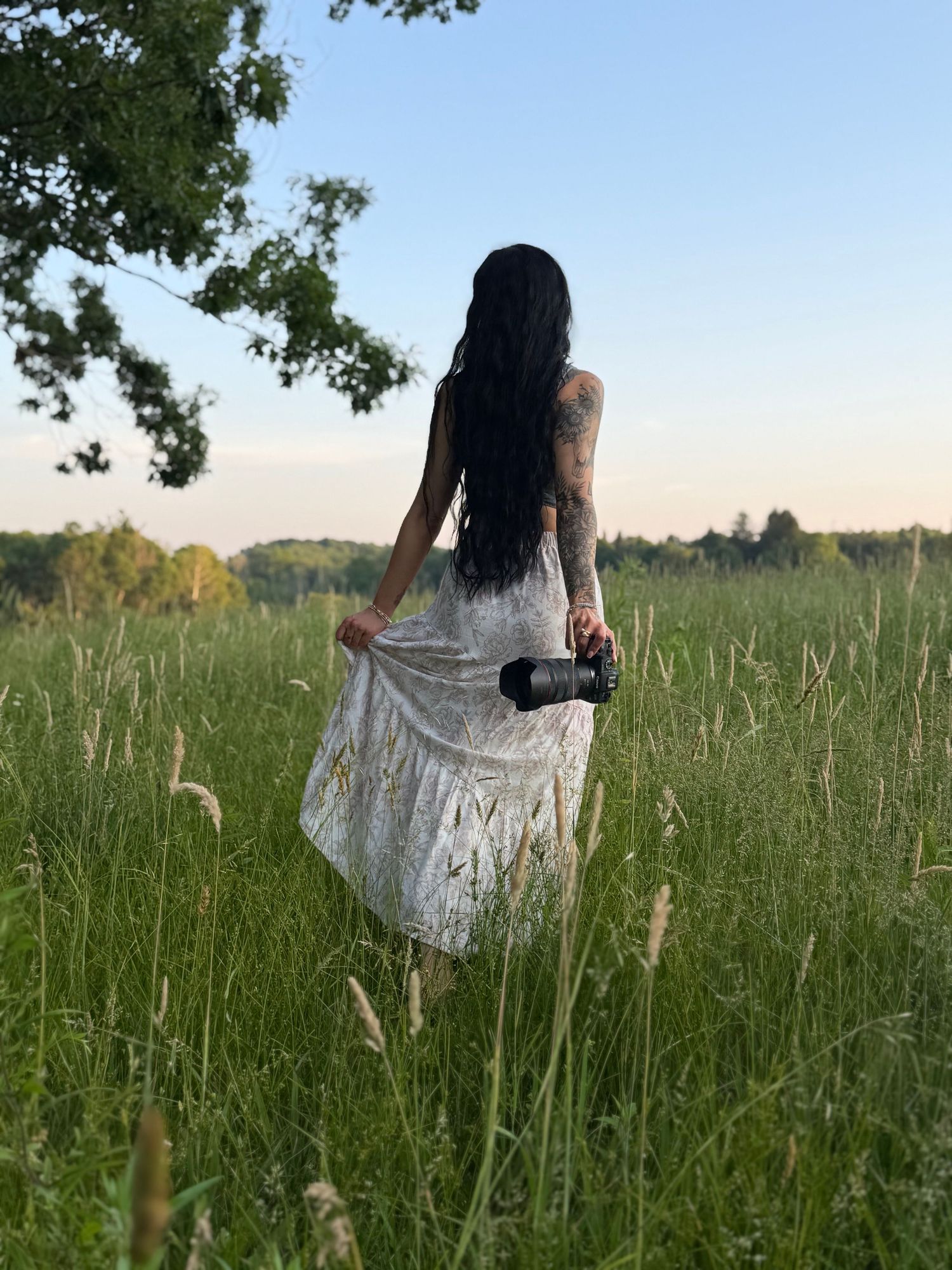 Woman in white dress holding camera stands in lush green field at dusk with long dark hair and tattoos.
