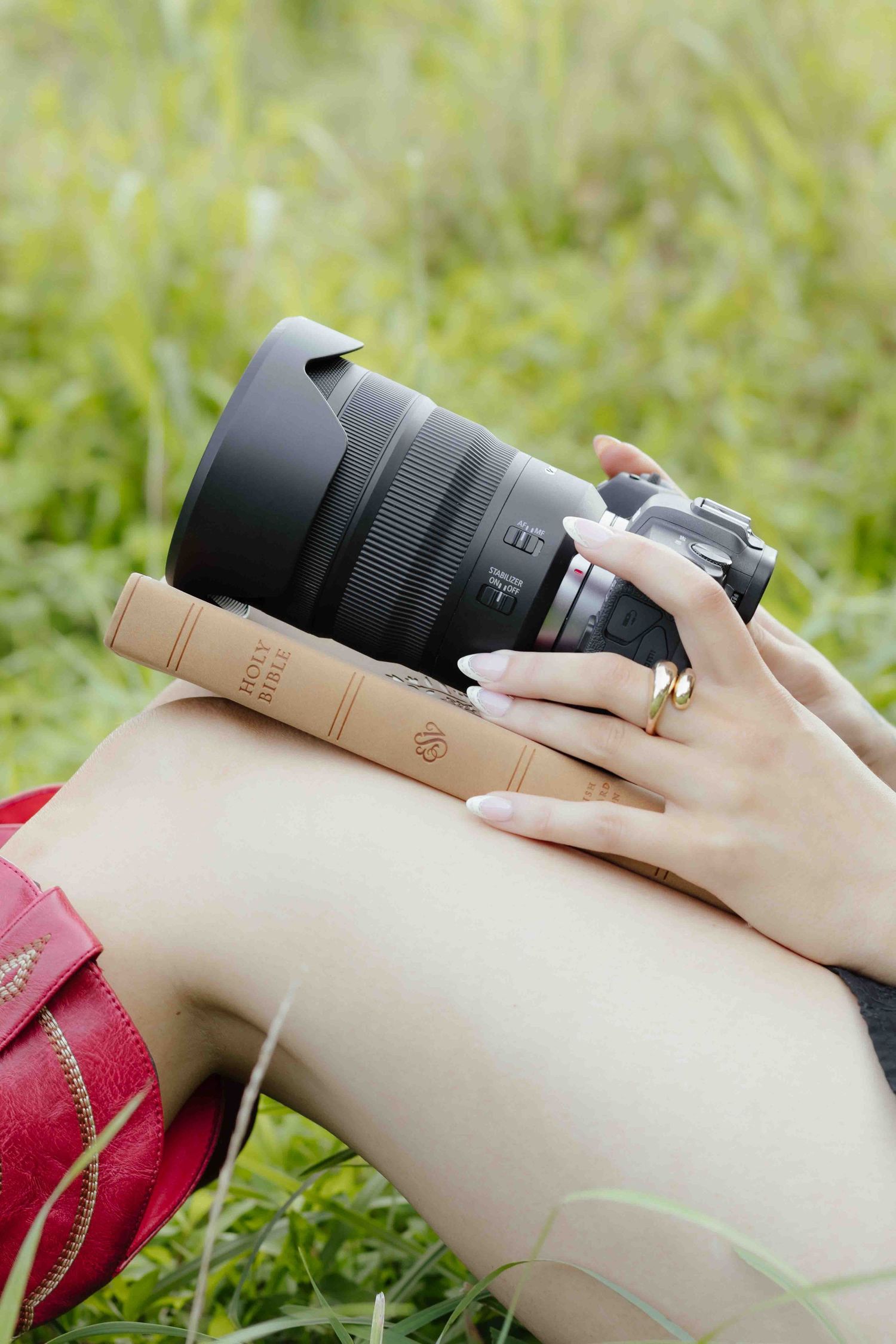 A woman holds a DSLR camera and vintage book on her knees while sitting in green grass, wearing a red skirt.