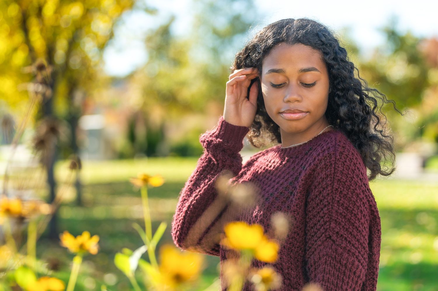 High school senior portrait of a young woman in a maroon sweater sitting in a field of yellow wildflowers.