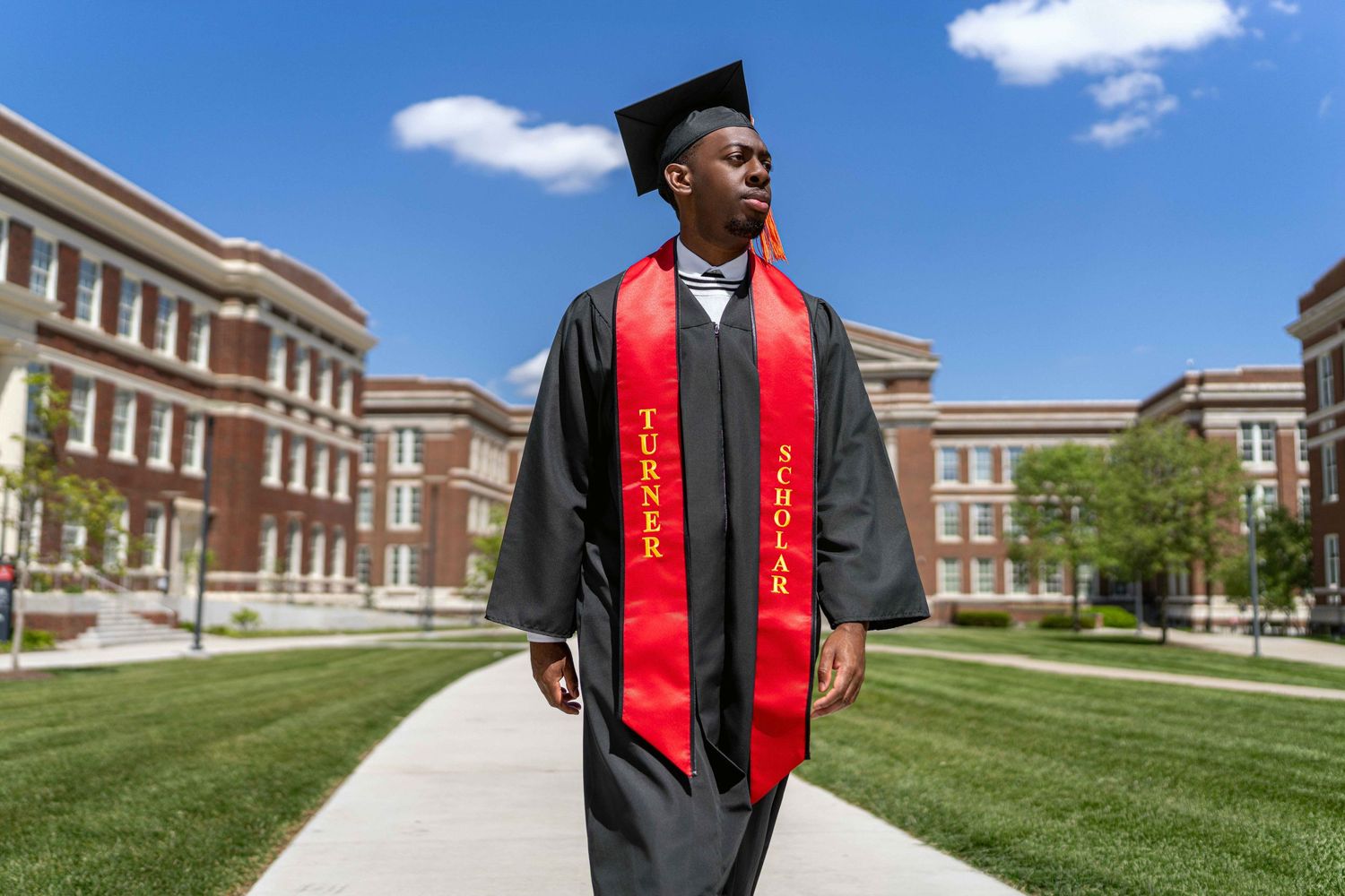 African American male in cap and gown with walking in front of a building looking away.